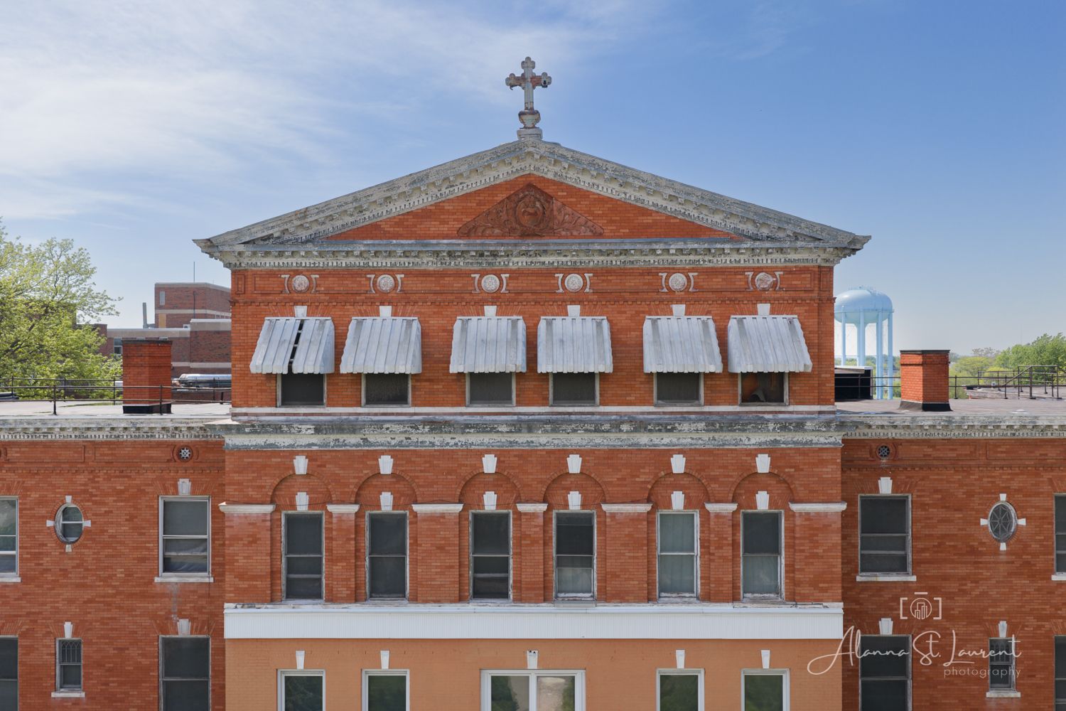Red brick church building with white-trimmed windows, decorative pediment, and cross on top against a clear blue sky.