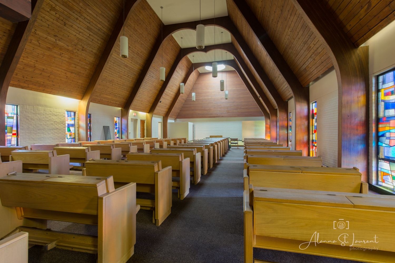 Modern church interior with wooden pews, vaulted ceiling, and colorful stained glass windows lining the walls.