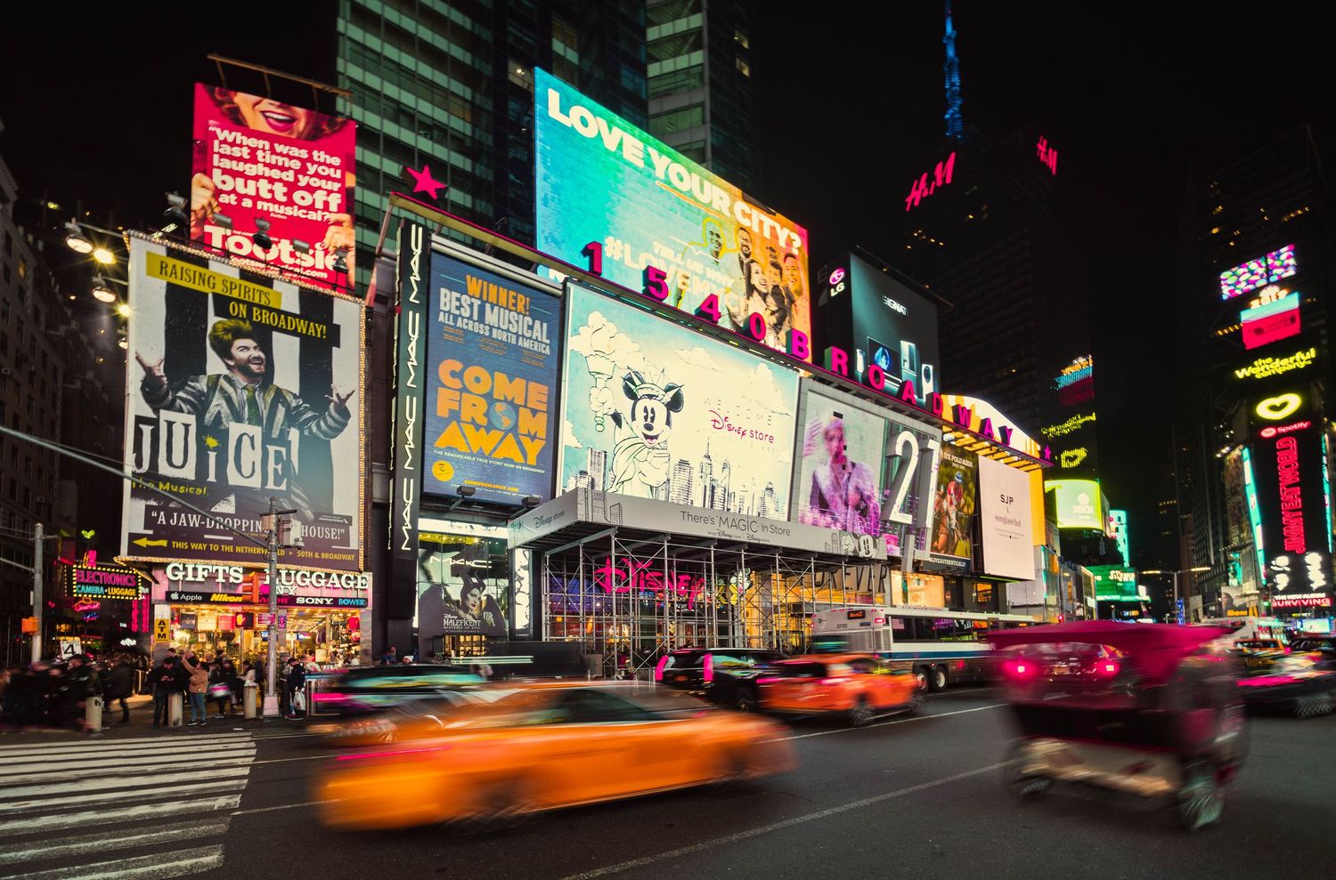 Vibrant nighttime cityscape of Times Square in Manhattan with bright neon signs and motion-blurred taxis creating light trails.