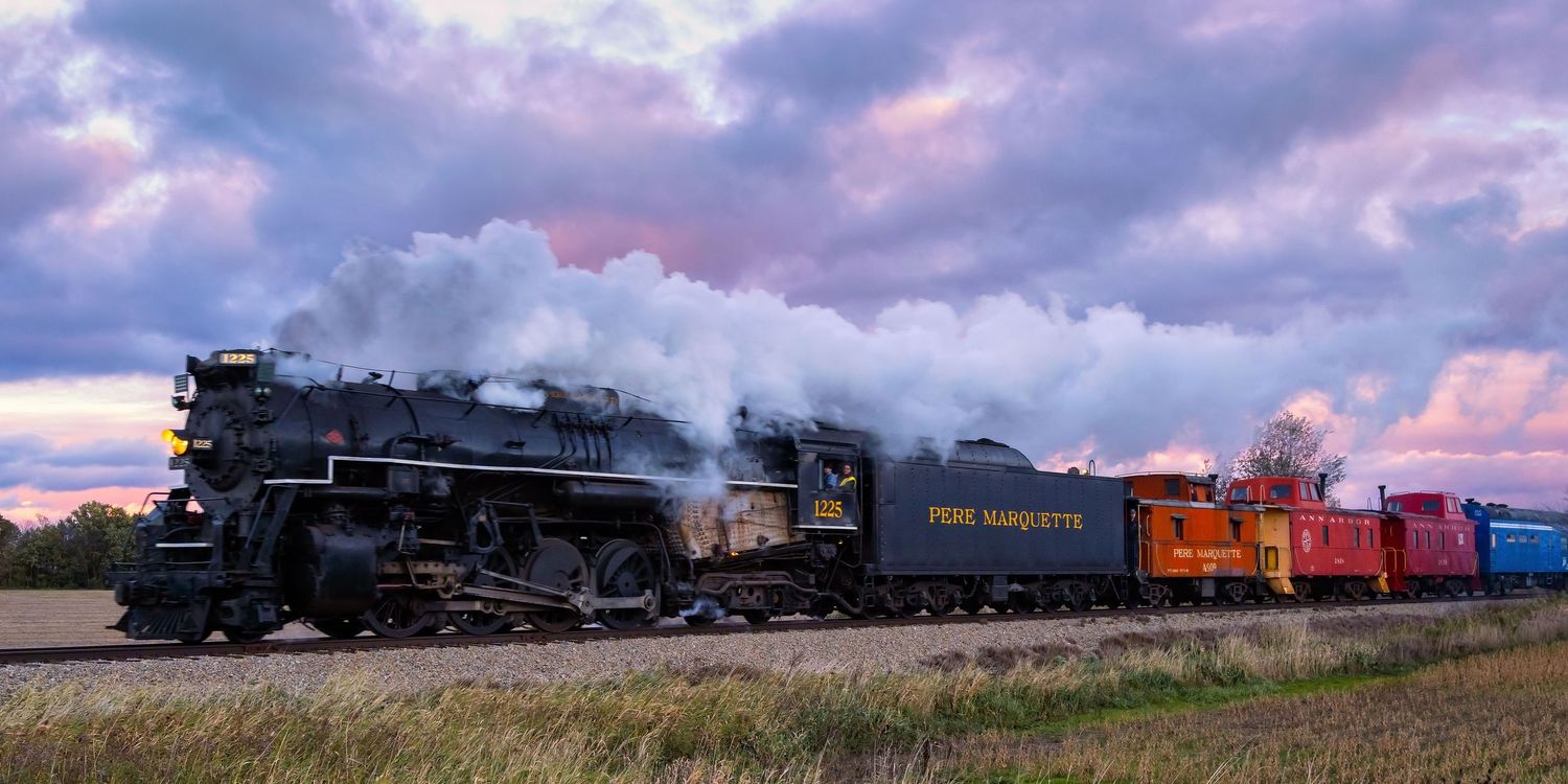 Pere Marquette 1225 steam locomotive pulls colorful passenger cars through countryside at sunset with dramatic purple clouds.