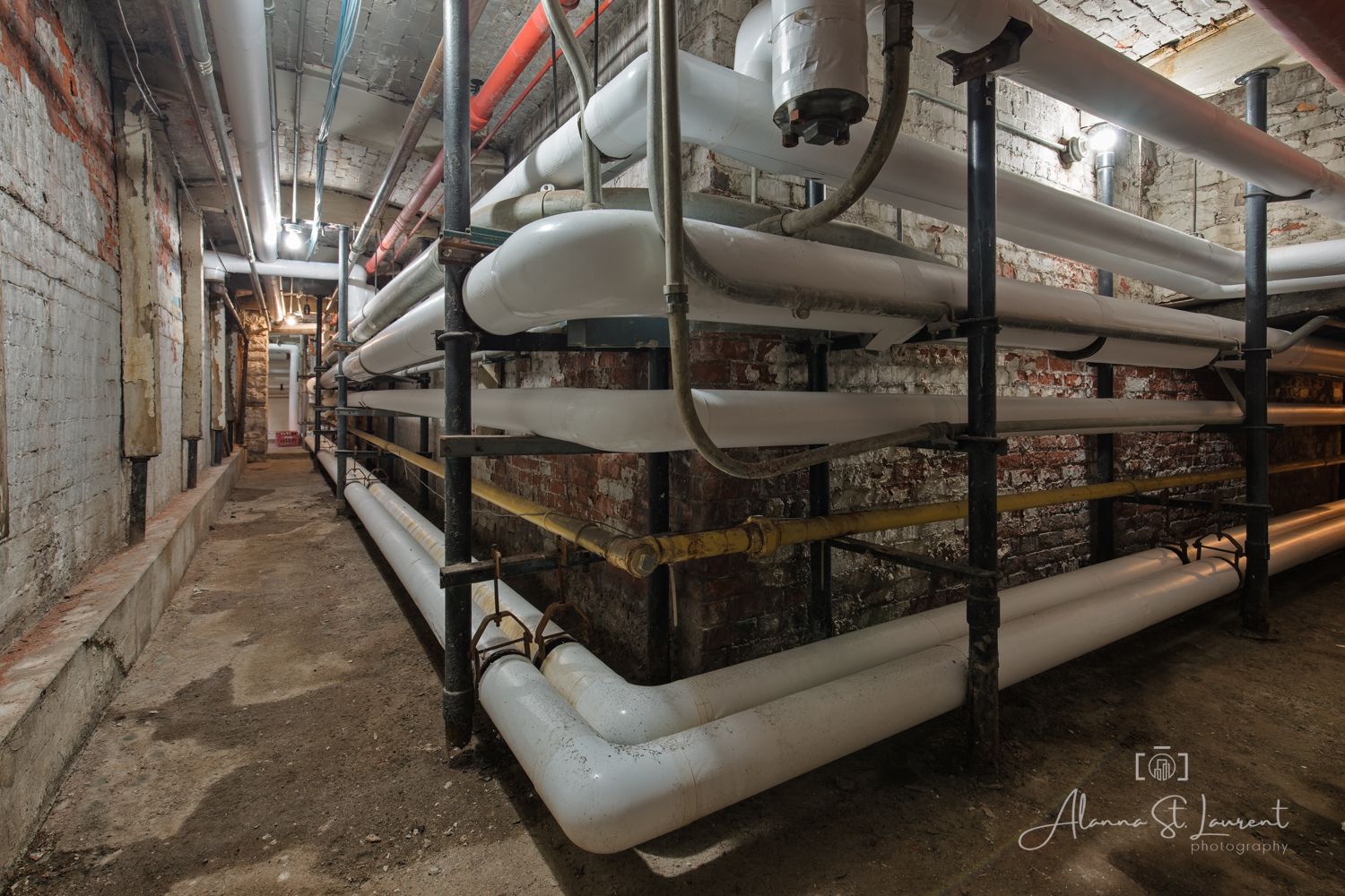 Abandoned industrial basement corridor with white pipes and metal shelving along graffiti-covered brick walls.