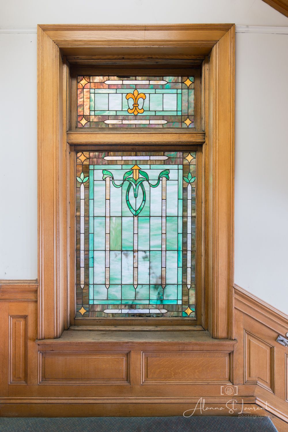 Ornate stained glass window with fleur-de-lis design in teal and gold tones, framed by warm wood paneling.