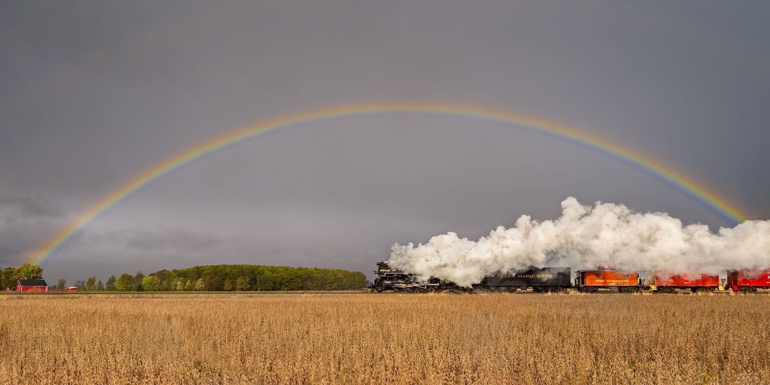 Pere Marquette 1225 steam train and colorful cars under a full rainbow arc stretching across stormy sky over rural farmland.