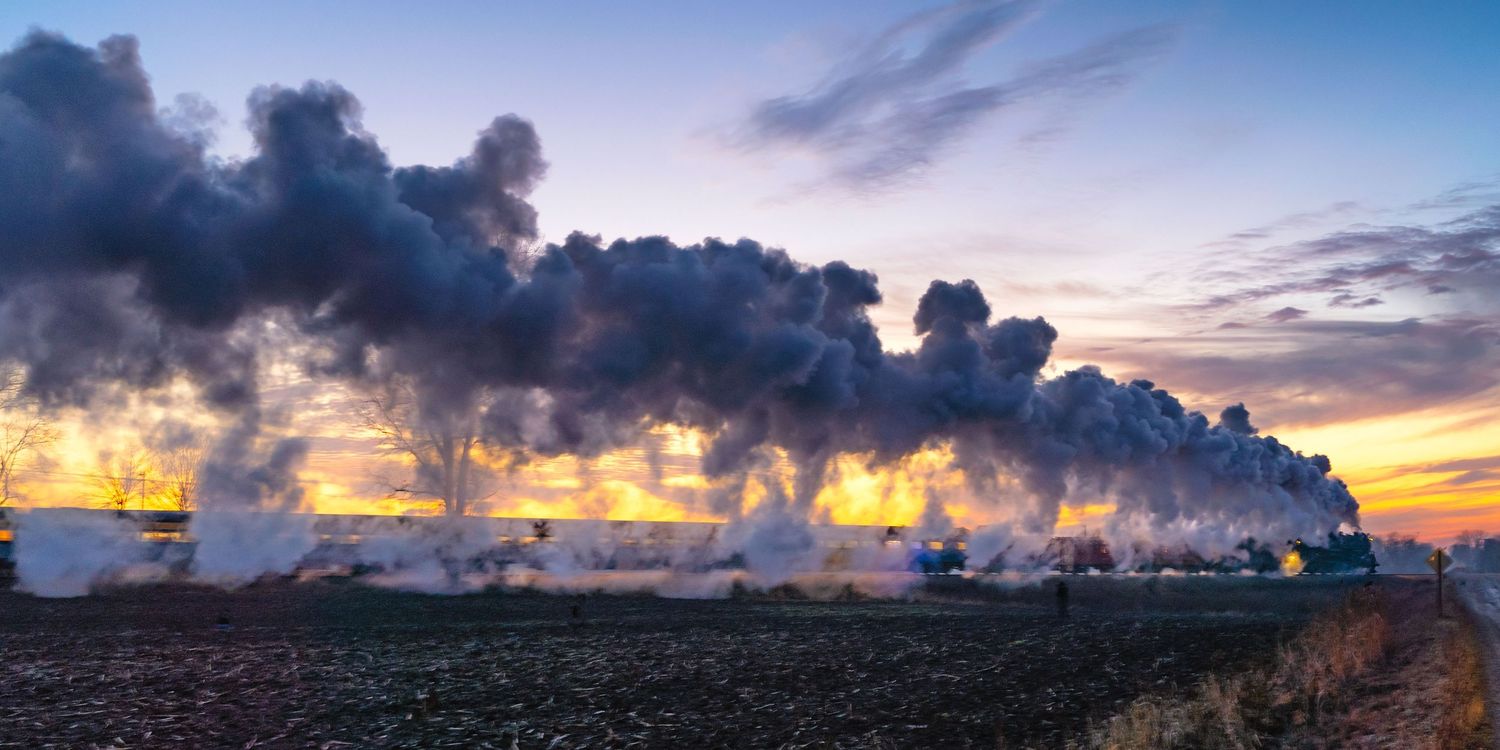Pere Marquette 1225 steam train creates dramatic silhouette against colorful sunset sky over rural landscape.