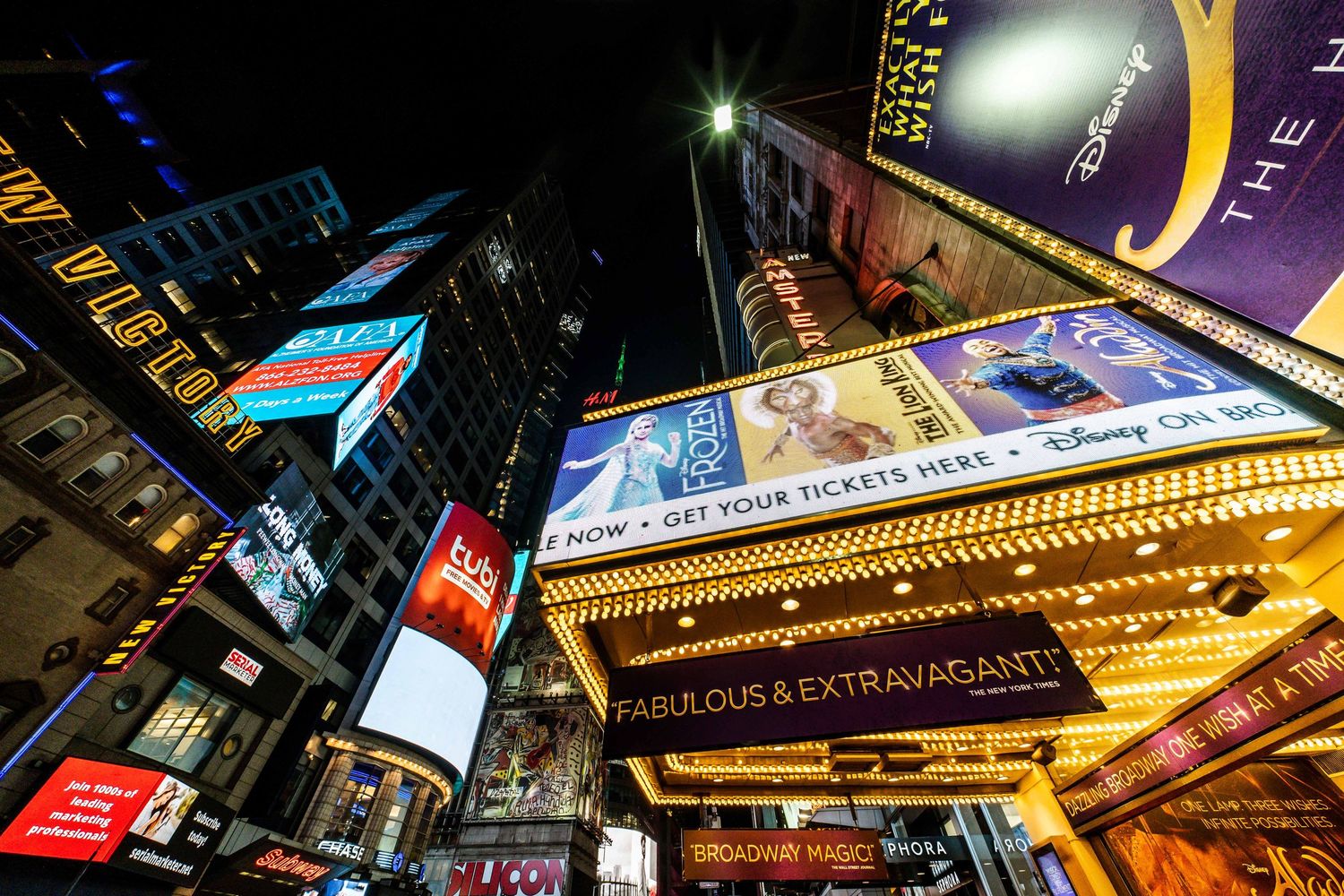 Illuminated Times Square billboards and theater marquees looking up at night in New York City.