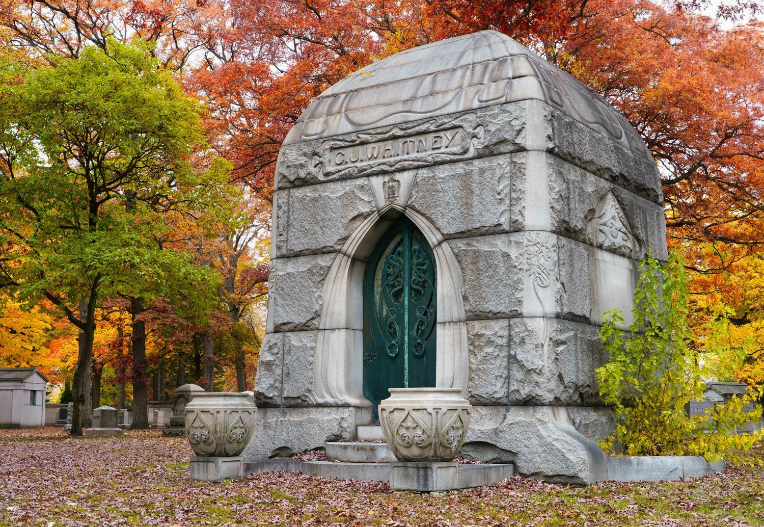CJ Whitney mausoleum with a green art deco door stands among vibrant orange and red autumn foliage in historic Woodlawn cemetery in Detroit.
