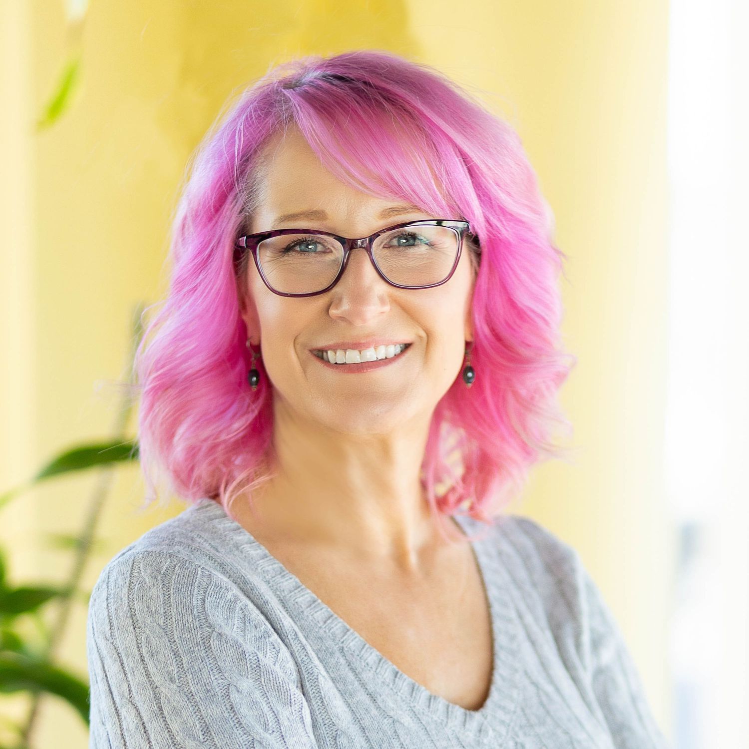 A portrait of Alanna St Laurent wearing a gray sweater with vibrant pink hair and glasses against a yellow background.