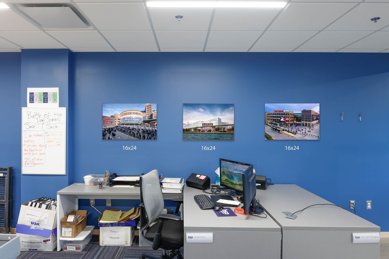A modern office space with blue walls features mounted photos and artwork along with white ceiling panels and lighting fixtures.