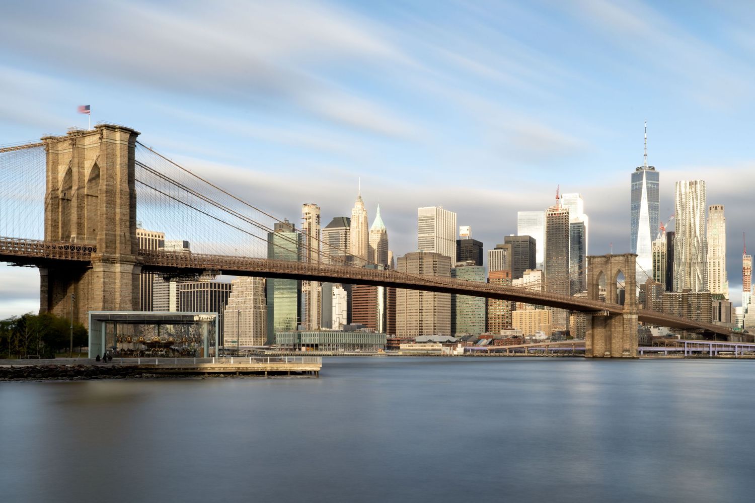 View of the Brooklyn Bridge and Manhattan skyline across calm East River waters with long exposure clouds.