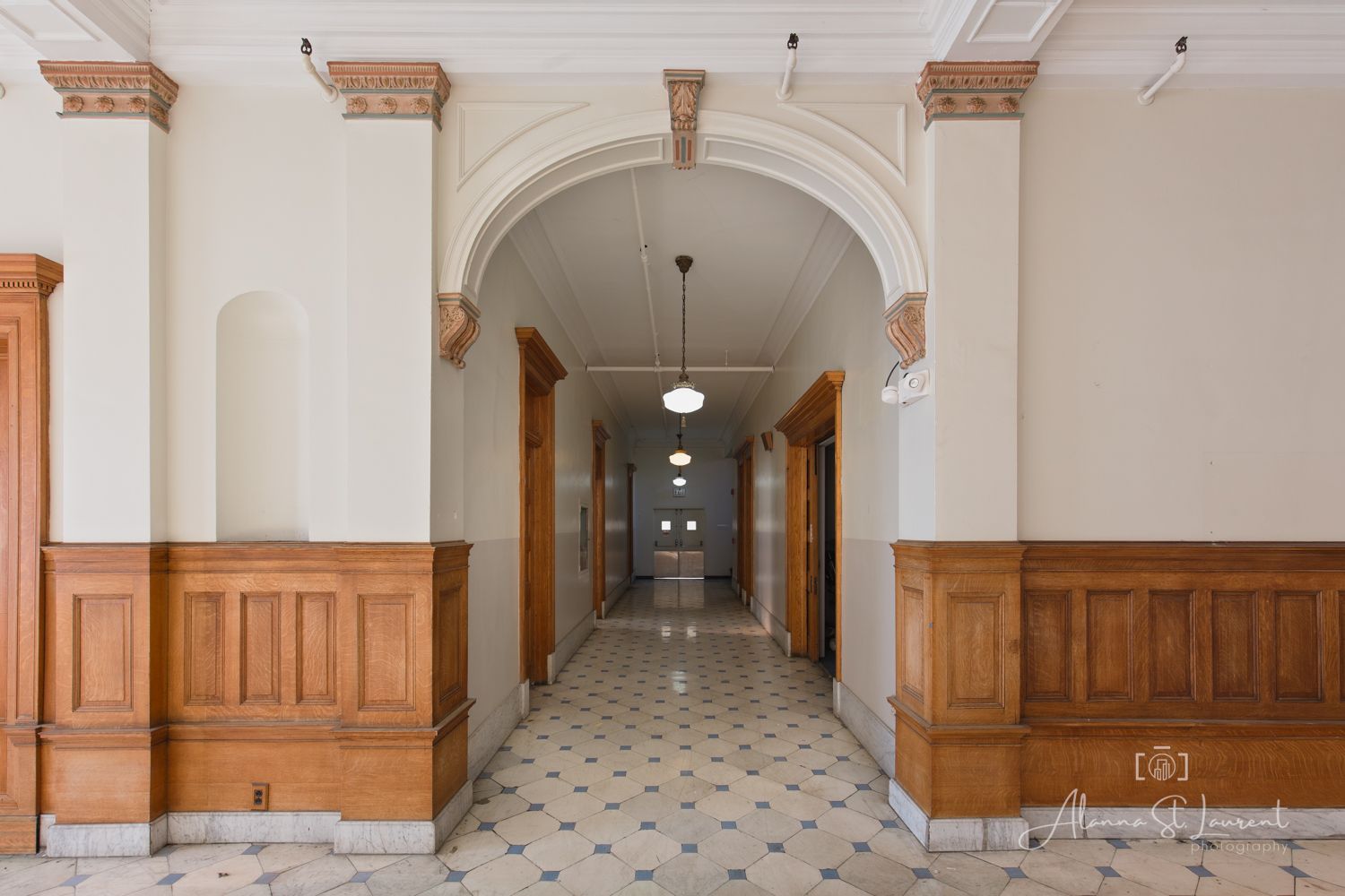Historic hallway with arched doorway, wainscoting, ornate columns, pendant lights, and checkered tile floors.