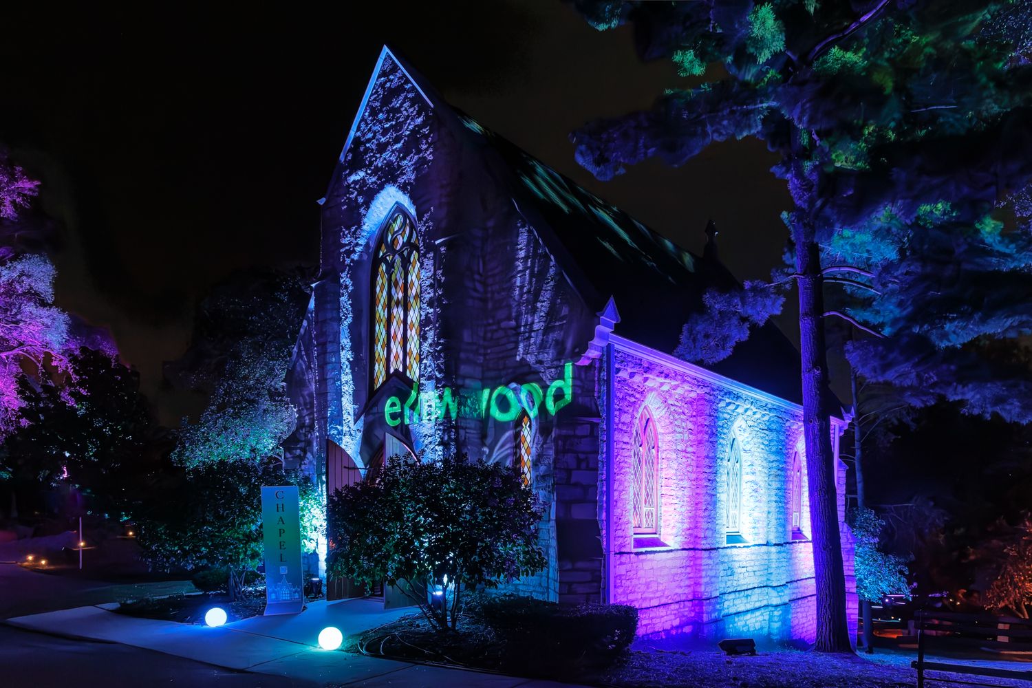 Historic chapel at Elmwood Cemetery in Detroit, Michigan bathed in colorful blue and purple lighting effects at night.