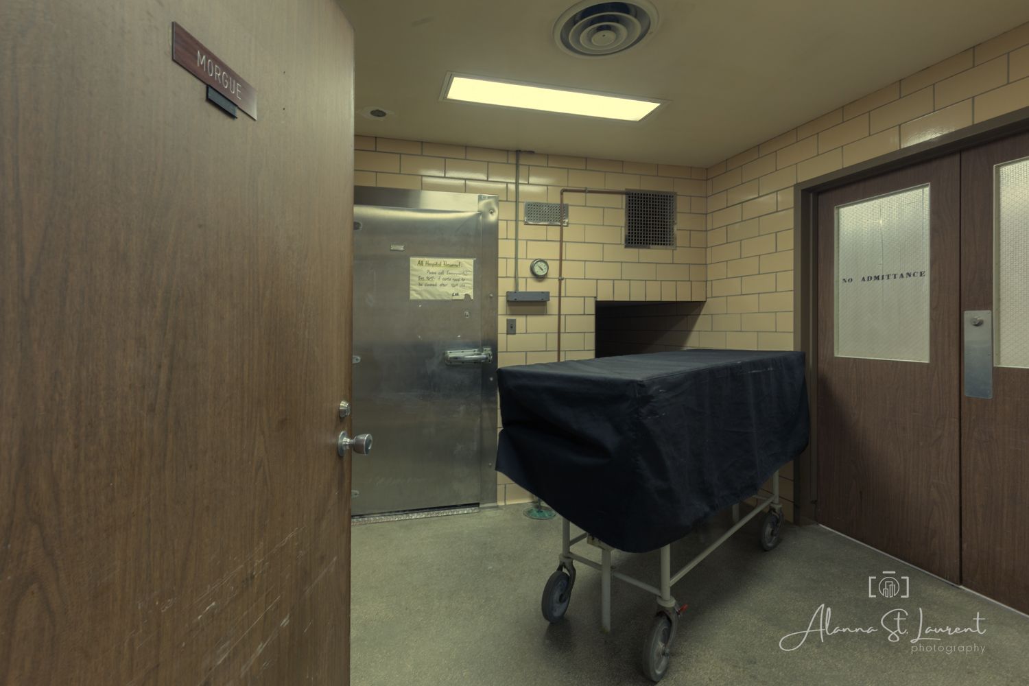 A dimly lit morgue room with a draped gurney on wheels, tiled walls, metal door, and institutional fluorescent lighting.