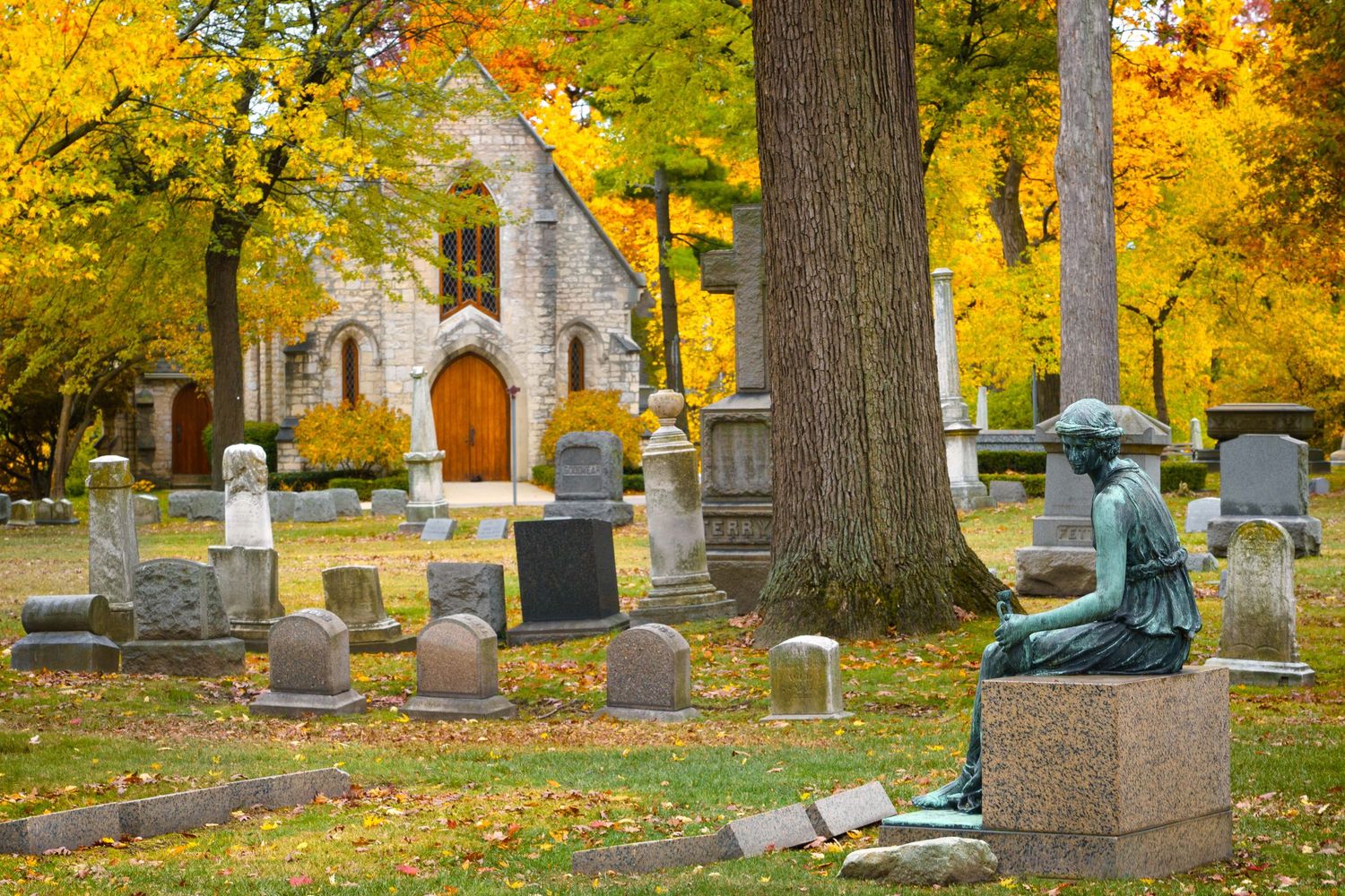 Historic Elmwood cemetery with weathered headstones, a statue and chapel in the background displaying golden autumn colors in Detroit, MI.