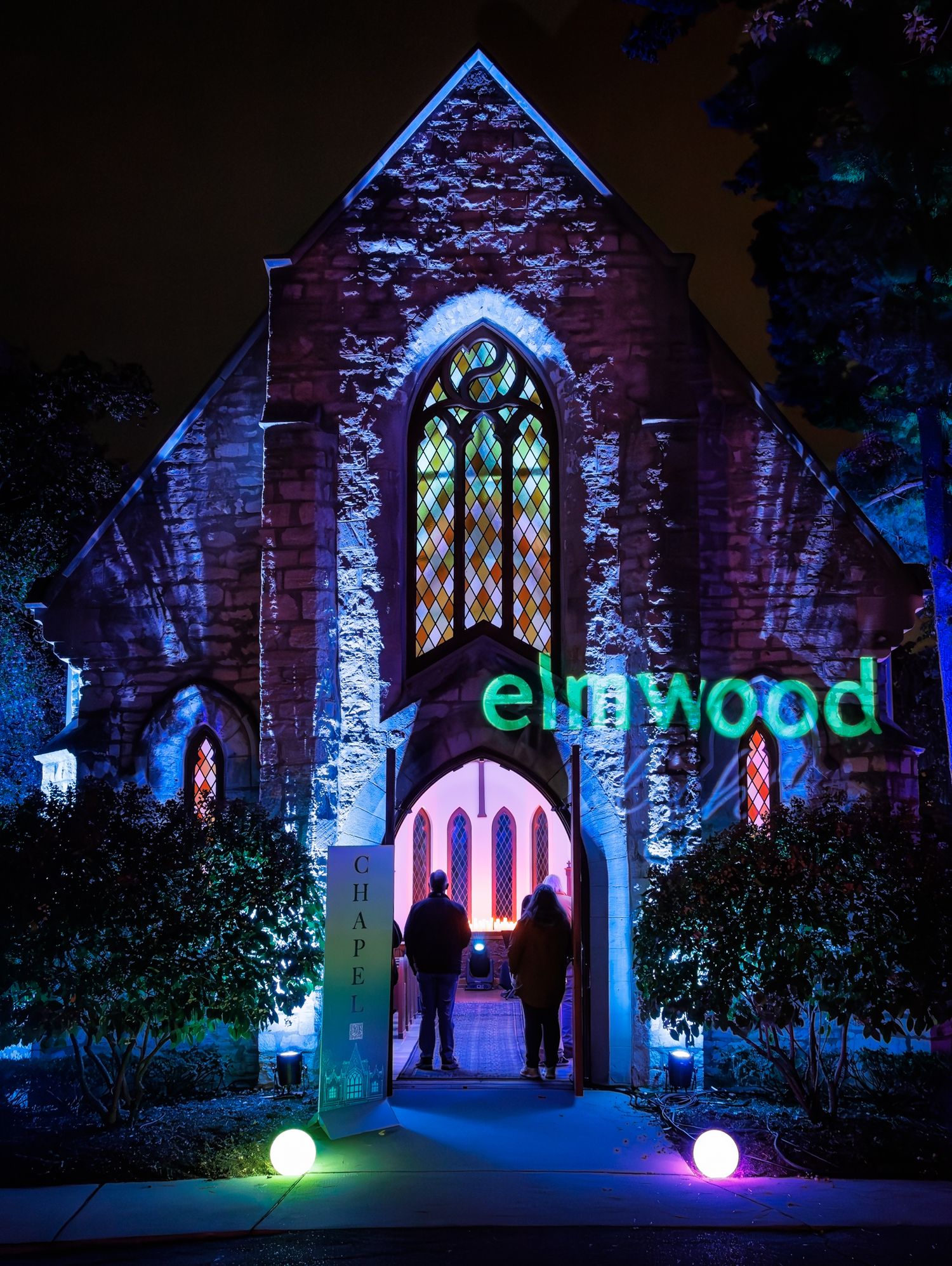 Historic chapel entrance at Elmwood Cemetery in Detroit Michigan illuminated with blue and purple lights highlighting stained glass windows.