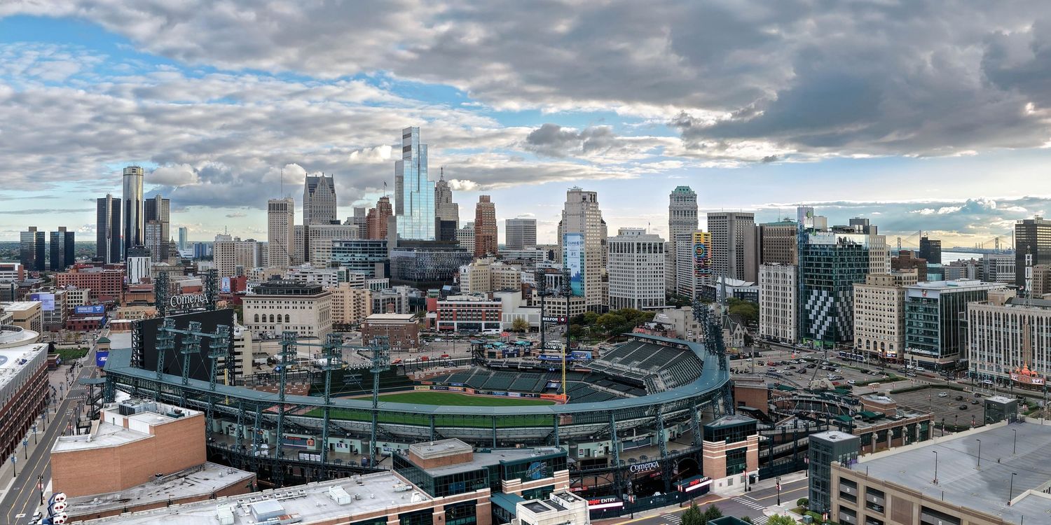 A panoramic aerial view of the Detroit city skyline with dramatic cloudy skies and modern buildings along the riverfront.