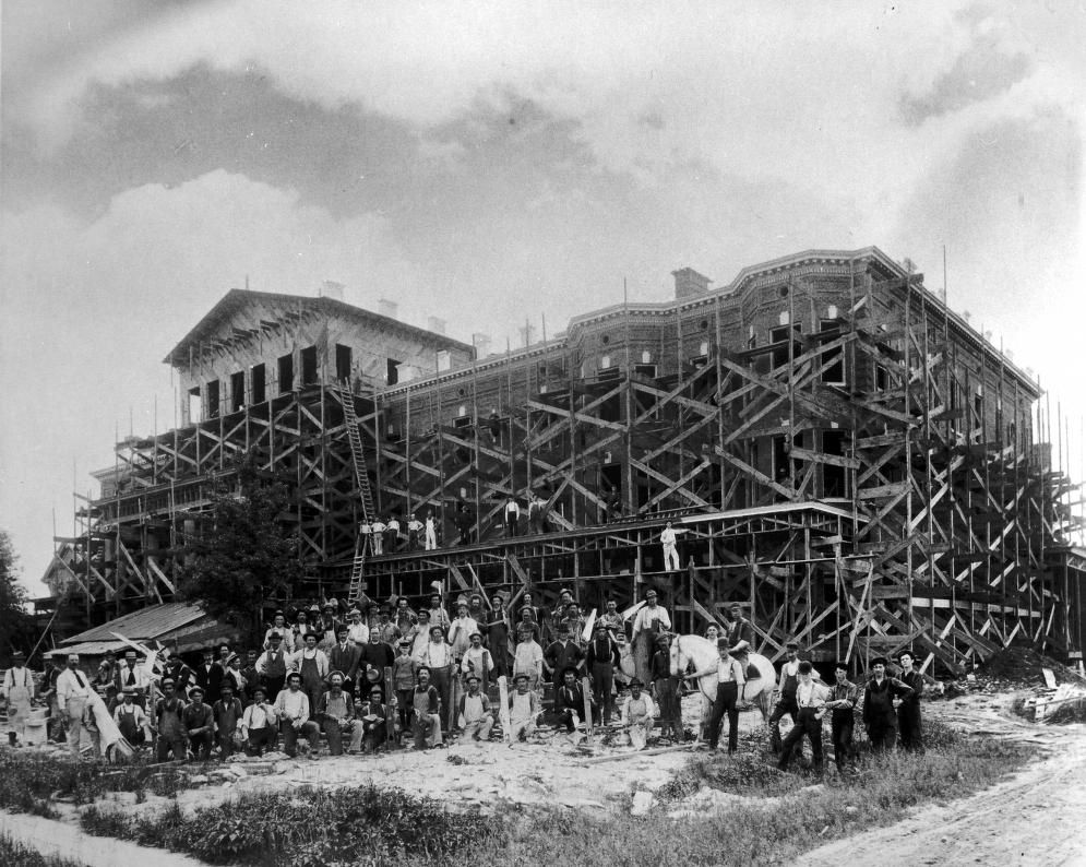 Historic black and white photo of a large building under construction with wooden scaffolding and workers gathered outside.