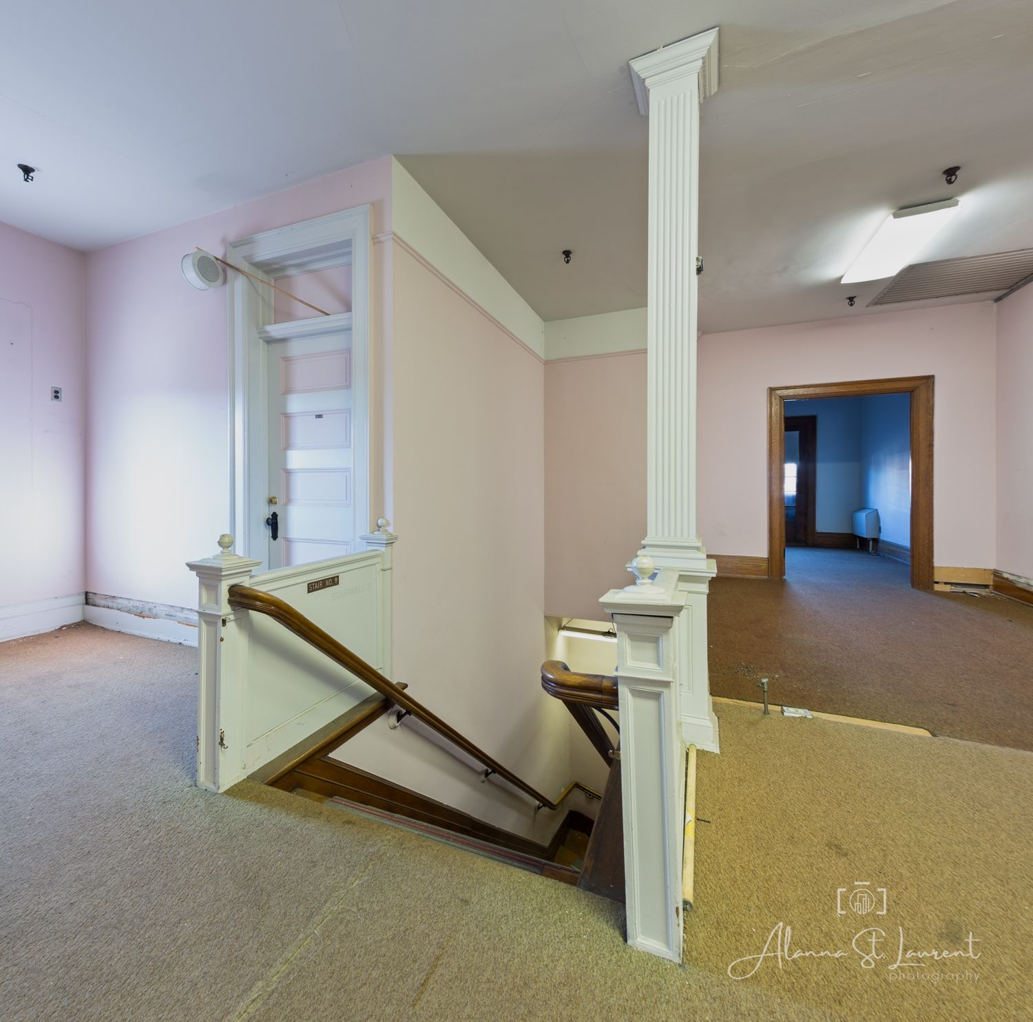 Empty room with pink walls, white columns, and a dismantled staircase railing on carpeted floor in an abandoned building.