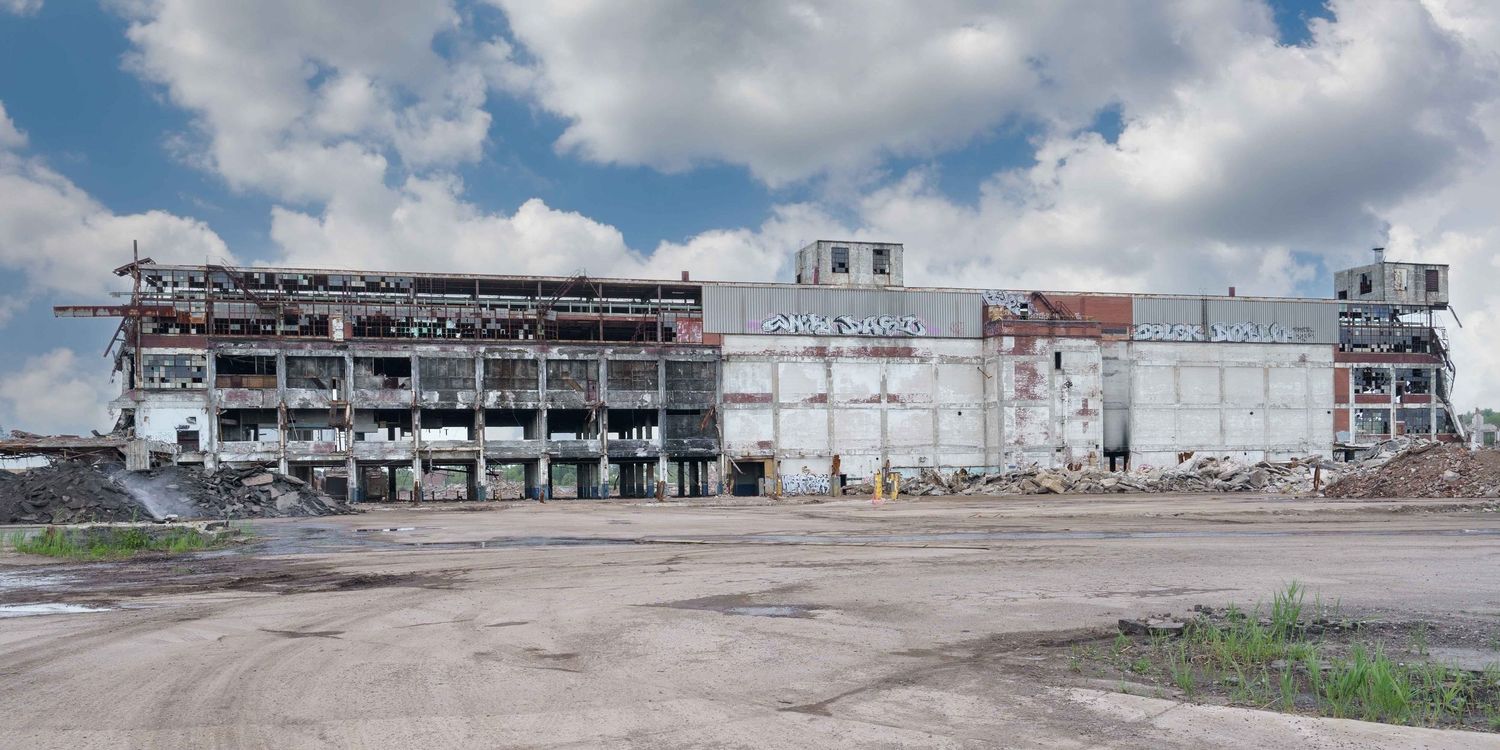 The front of the abandoned Cadillac Stamping plant in Detroit during demolition under a bright blue sky and white clouds.