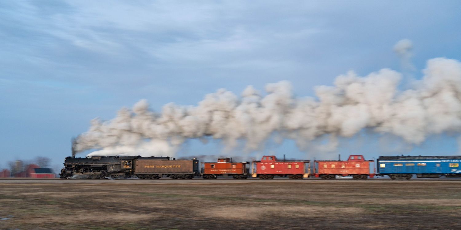 Pere Marquette 1225 steam locomotive leads colorful train cars across open farmland under a cloudy sky with trailing steam.