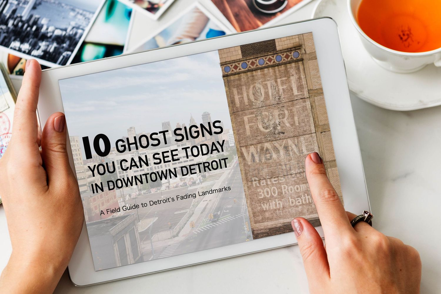 Hands holding a tablet displaying a guide to 10 ghost signs in downtown Detroit, with a cup of tea nearby.