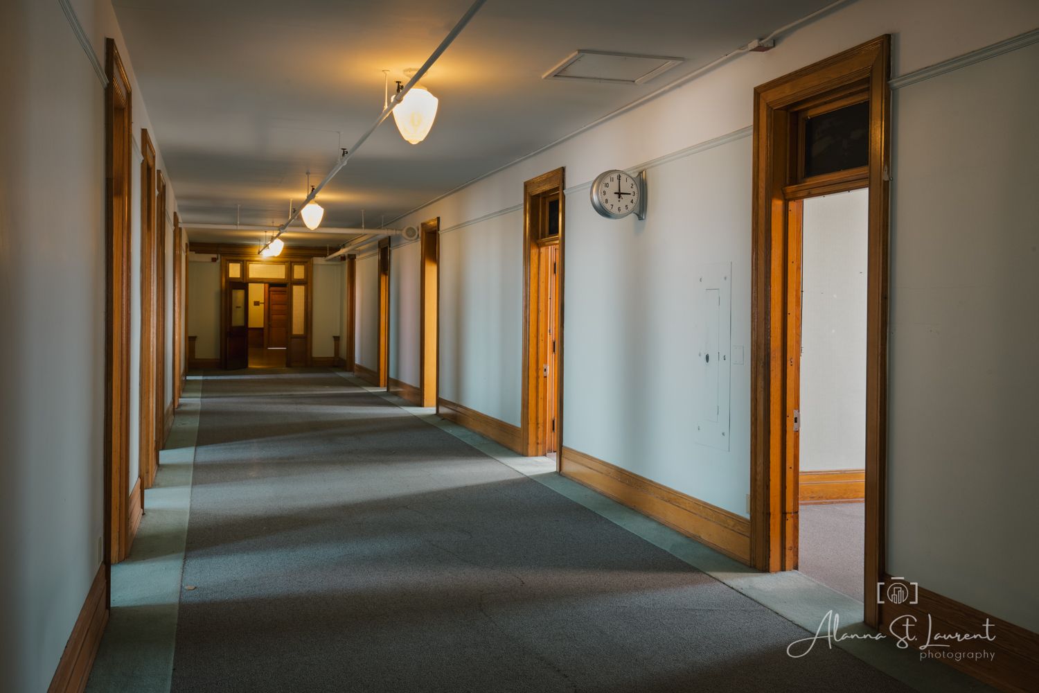 Long empty hallway with wooden doors, gray carpet, and ceiling lights in a vintage institutional building.