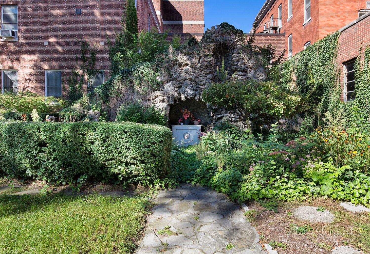 Lush garden grotto with stone pathway, ivy-covered walls, and dense greenery beside a brick building on a sunny day.