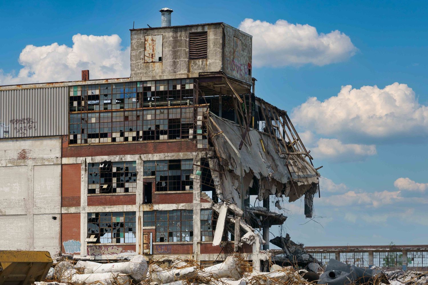 A phase of demolition of the Cadillac Stamping plant in Detroit with exposed steel beams and broken walls against blue sky and white clouds.