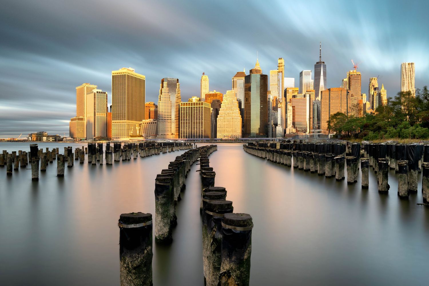 Golden hour cityscape of Manhattan with wooden pier posts from Brooklyn with long exposure clouds.