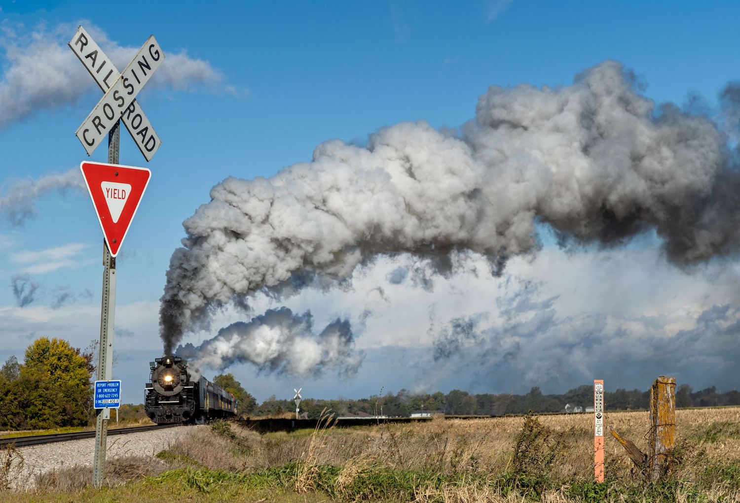Pere Marquette 1225 steam train passes railroad crossing sign while releasing thick clouds of steam.