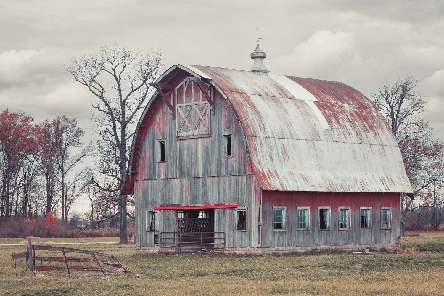 A weathered red barn with curved metal roof in a rural setting with a small fence in the foreground in rural Indiana.