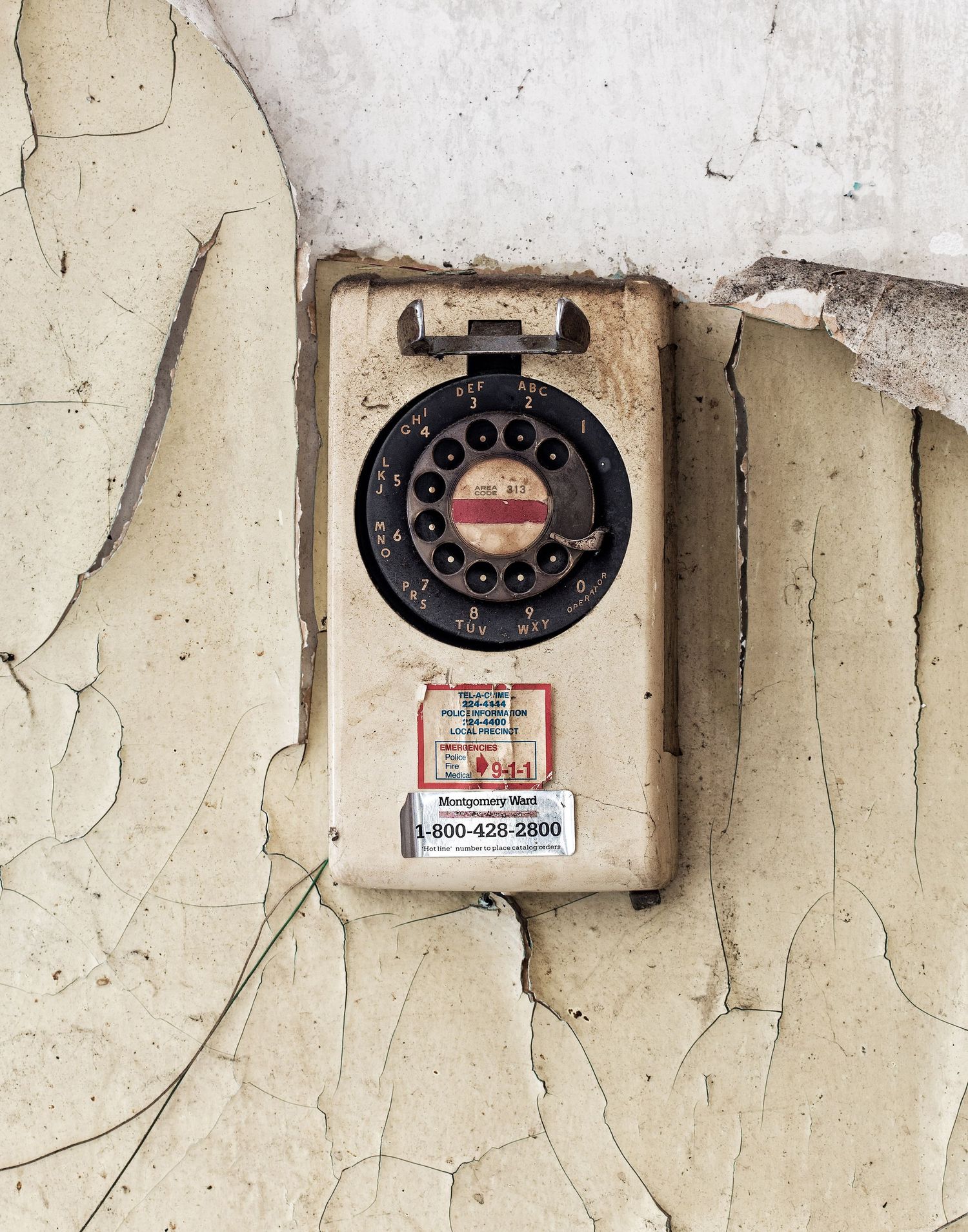 An old rotary phone mounted on a deteriorating beige painted wall in a kitchen of an abandoned home in Detroit, Michigan.