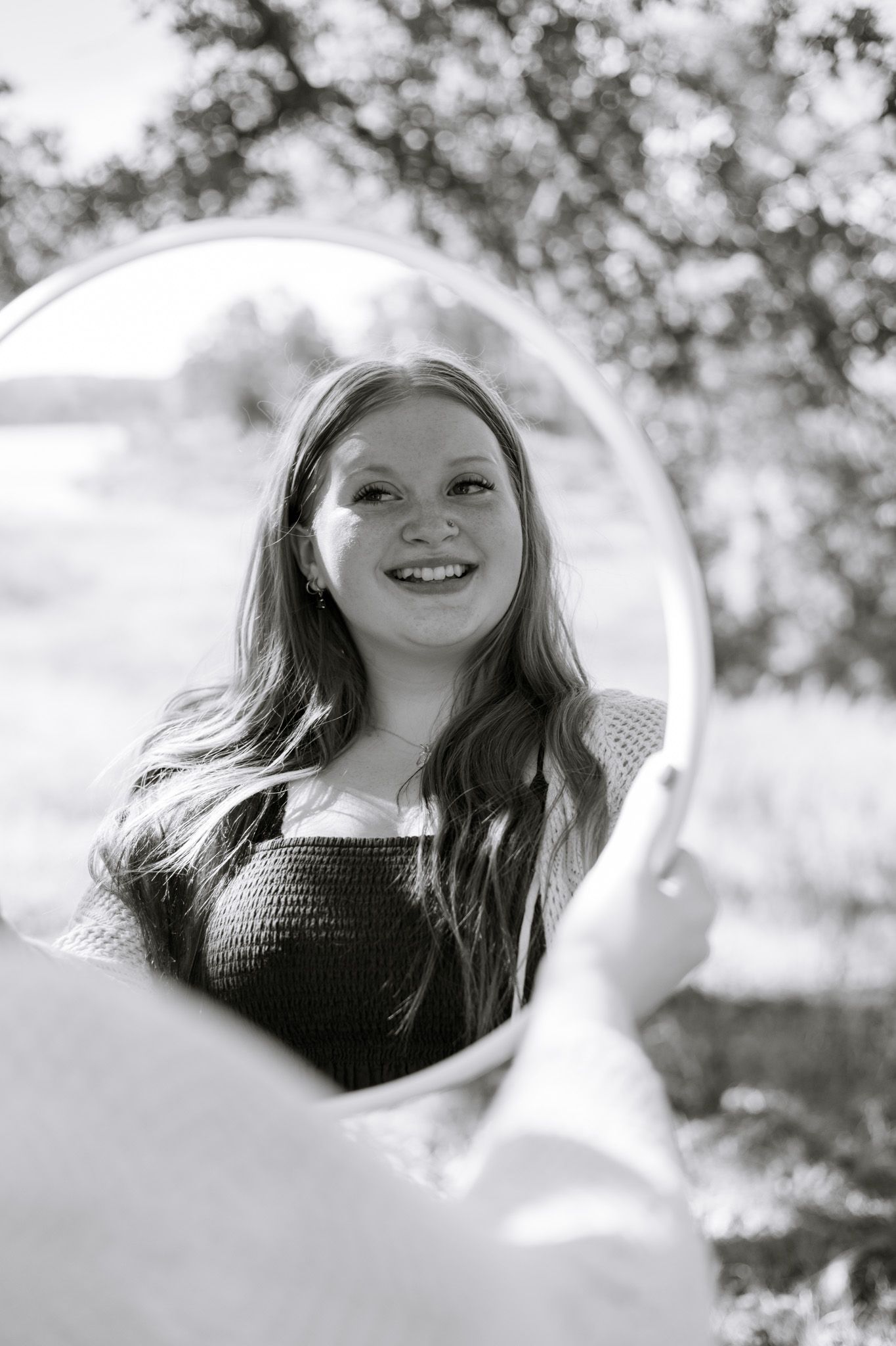 A young adult girl holds up a mirror and smiles at herself in it. The image is in black and white.