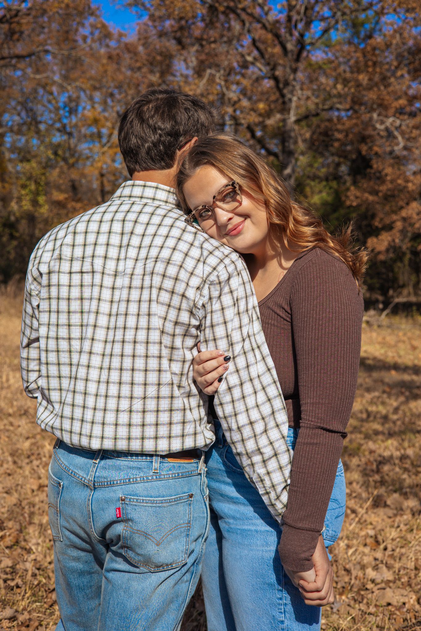 A young adult girl hugging her boyfriend. She is facing the camera and lays her head on his shoulder while he is faced behind her.