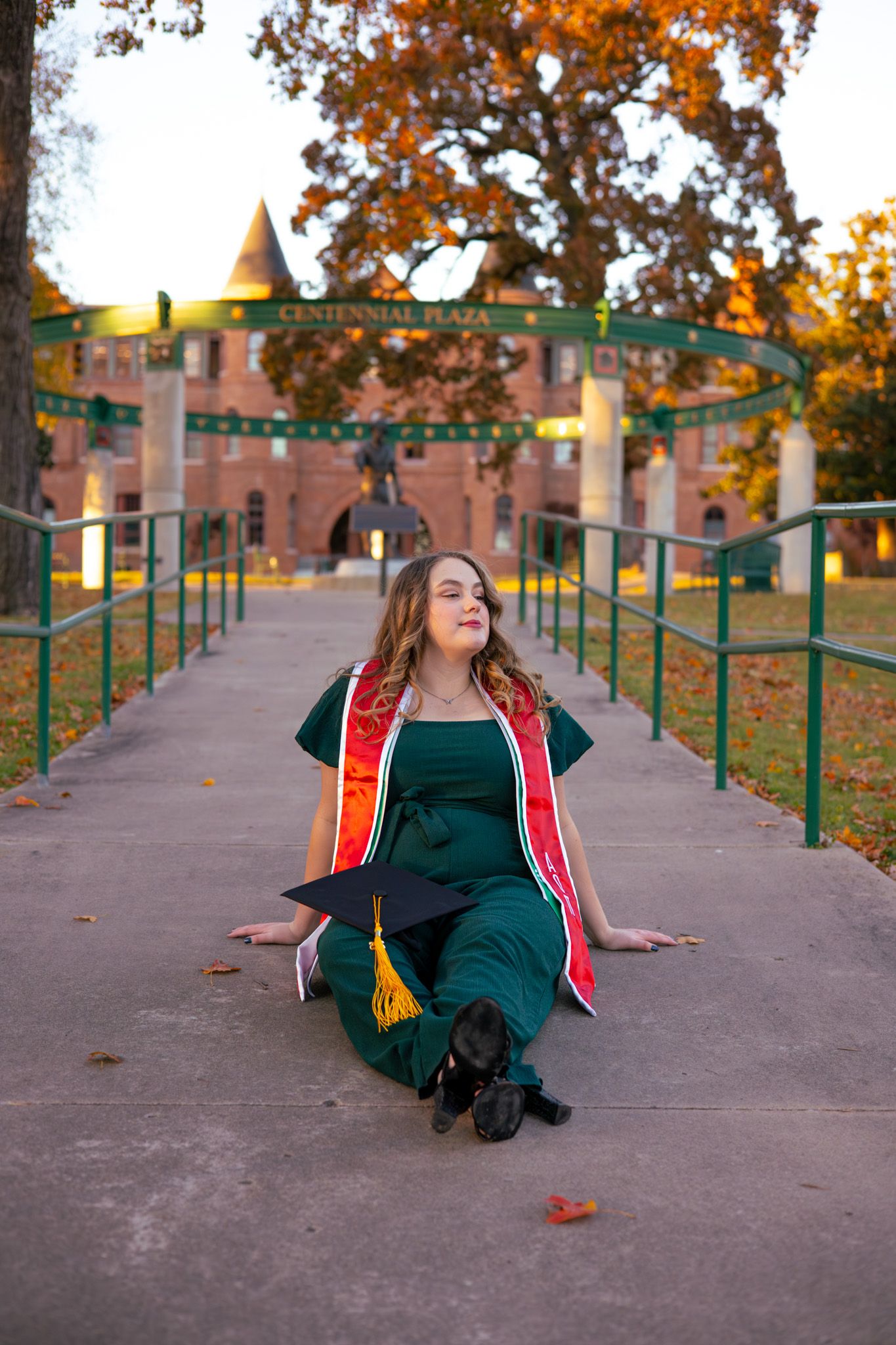 A young adult girl in her senior regalia. She is sitting up on the sidewalk facing away from the camera.