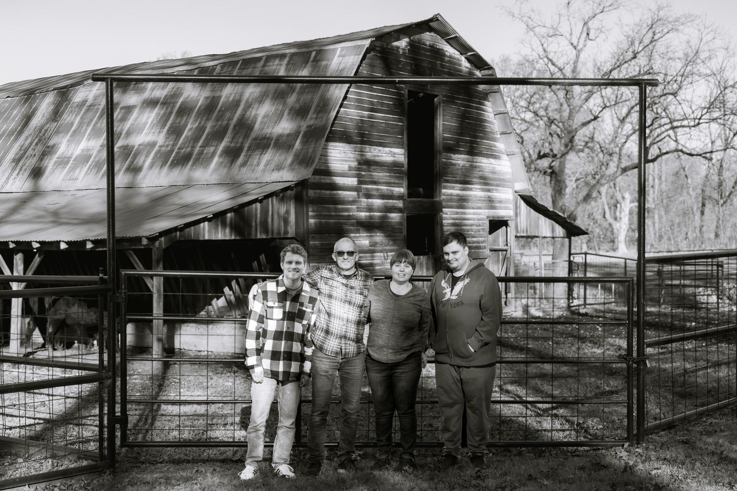 A family of four is placed in front of an old barn and black fence. The image is underexposed and in black and white.