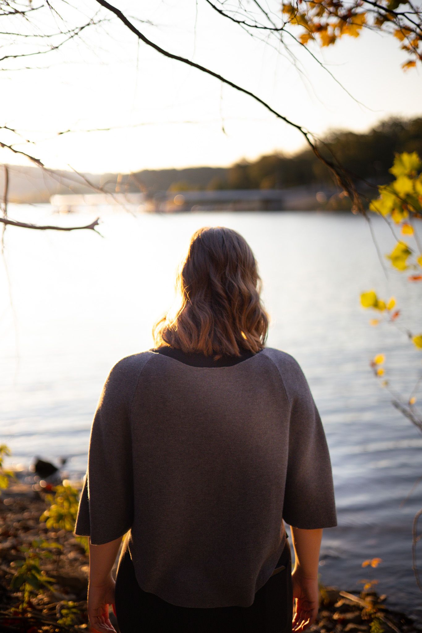 A young adult girl faces away from the camera and looks into a lake and a sunset.