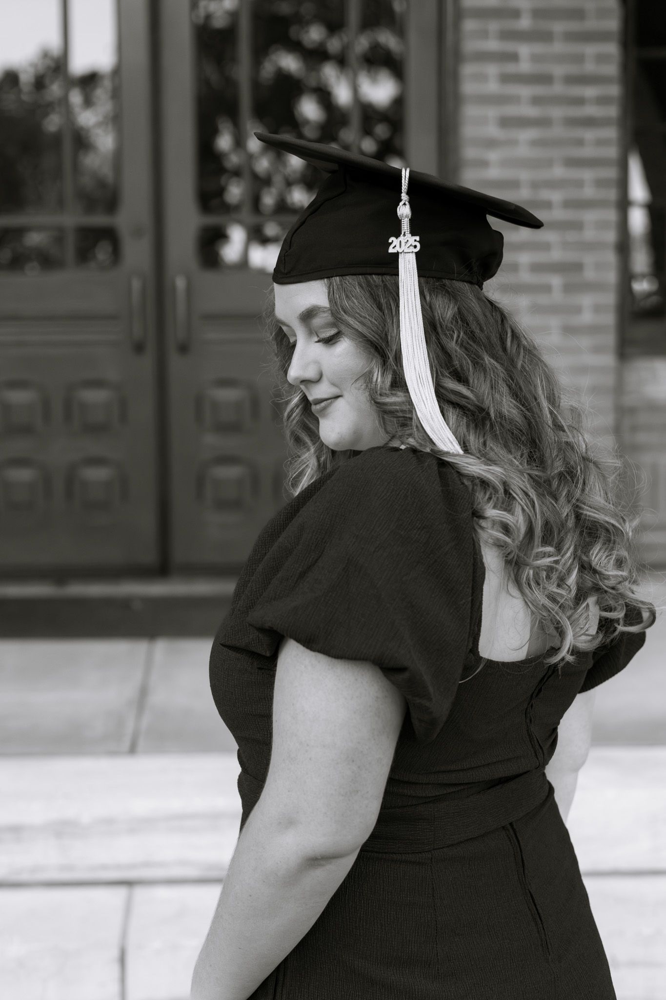 A young adult woman wearing a graduation cap and looks down at the ground with her eyes closed. The image is in black and white.