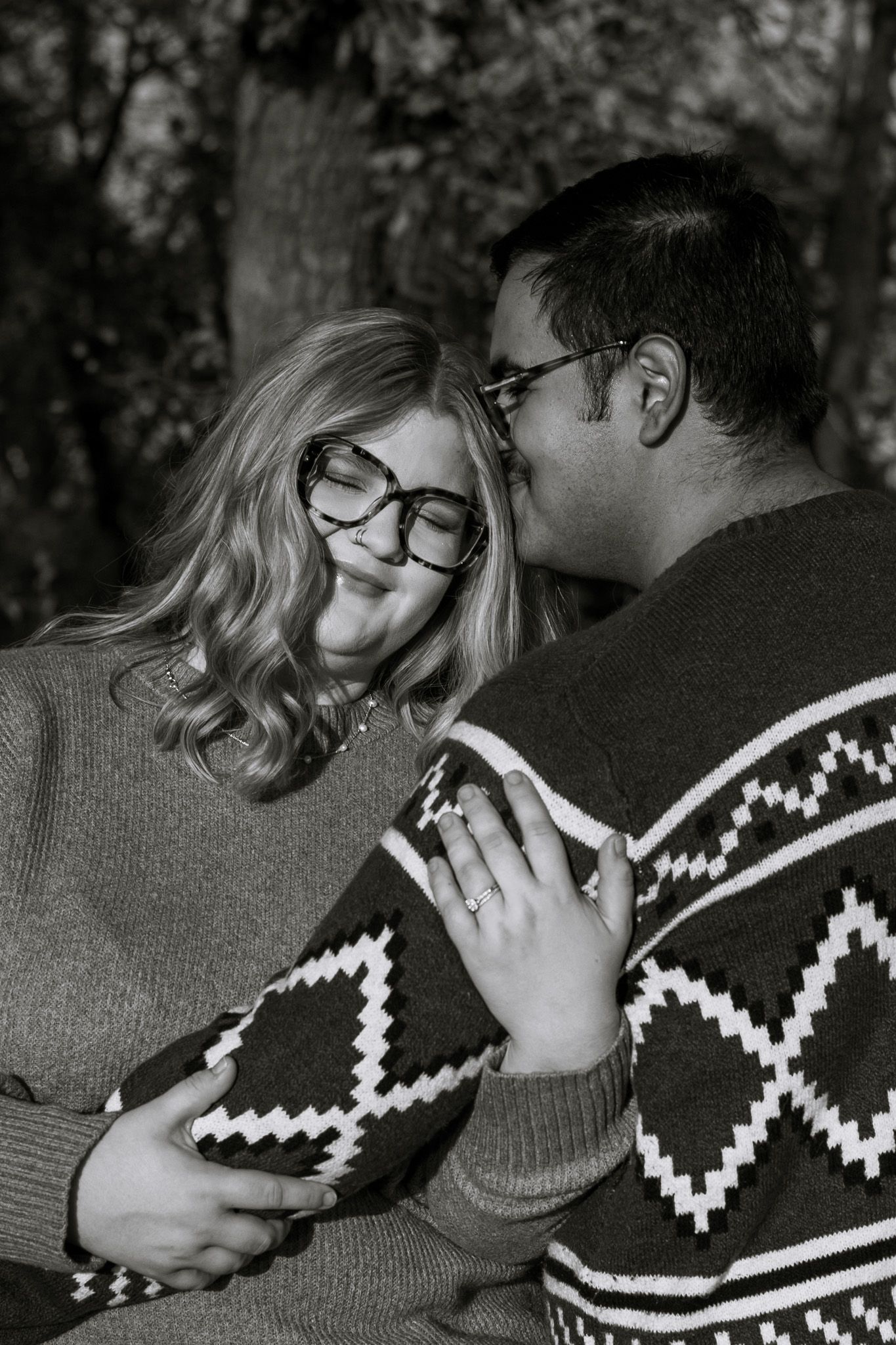 A young adult couple embraces closely. The male is kissing the female and she smiles with her eyes closed. The image is in black and white.