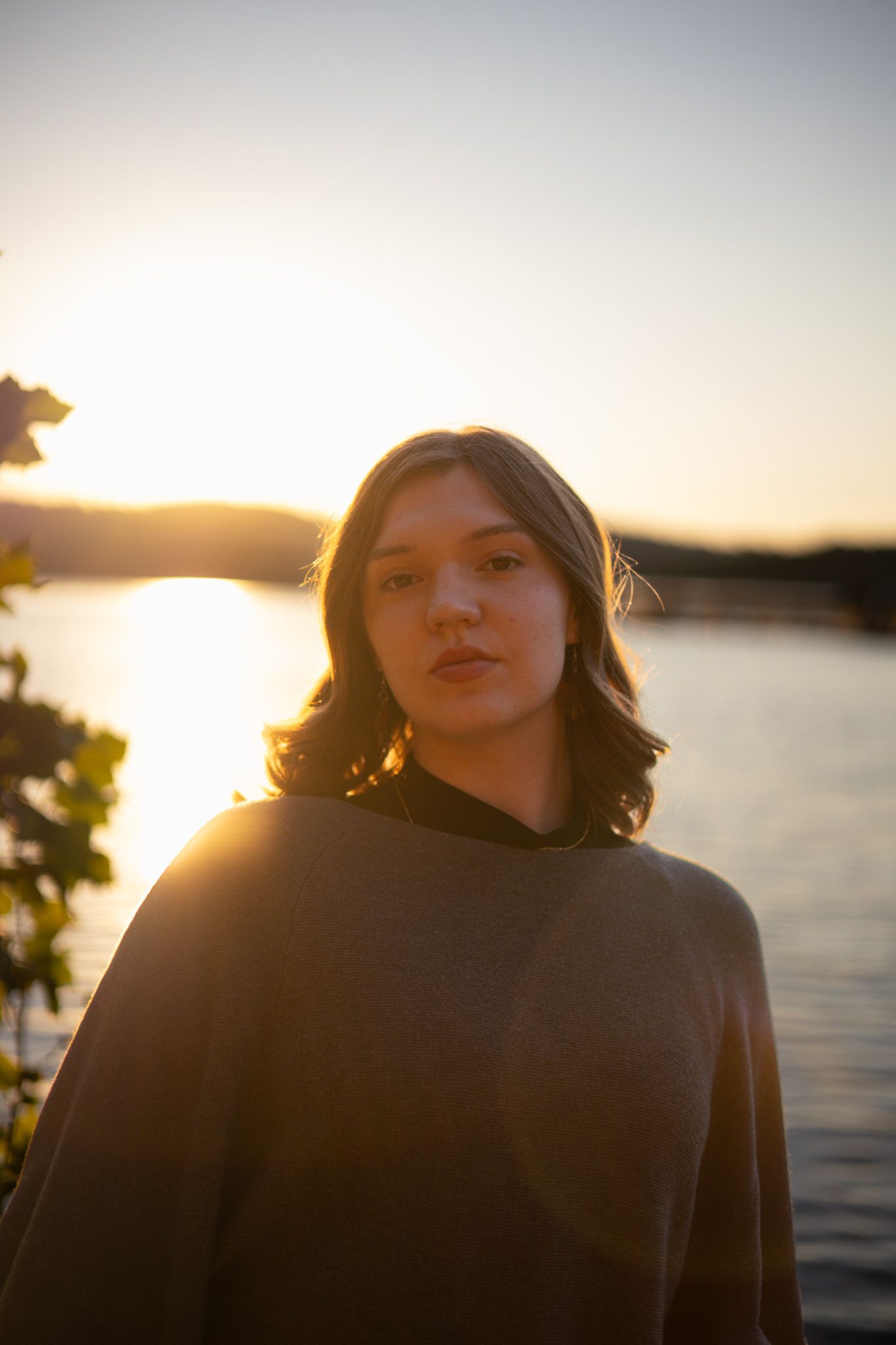 A young adult girl faces the camera in front of a lake with a sunset. She is not smiling for a dramatic image effect.