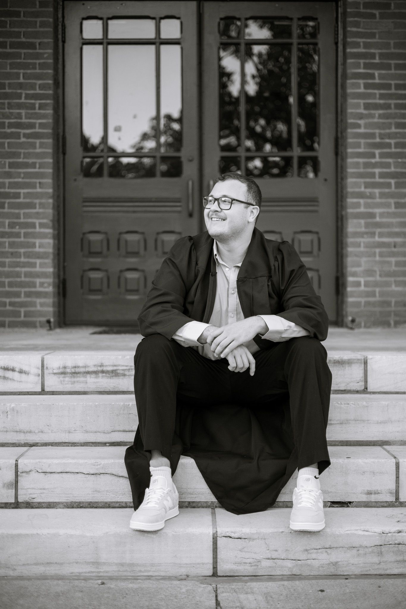 A young adult boy wearing a graduation gown. He sits on the stairs and looks off with a smile. The image is in black and white.