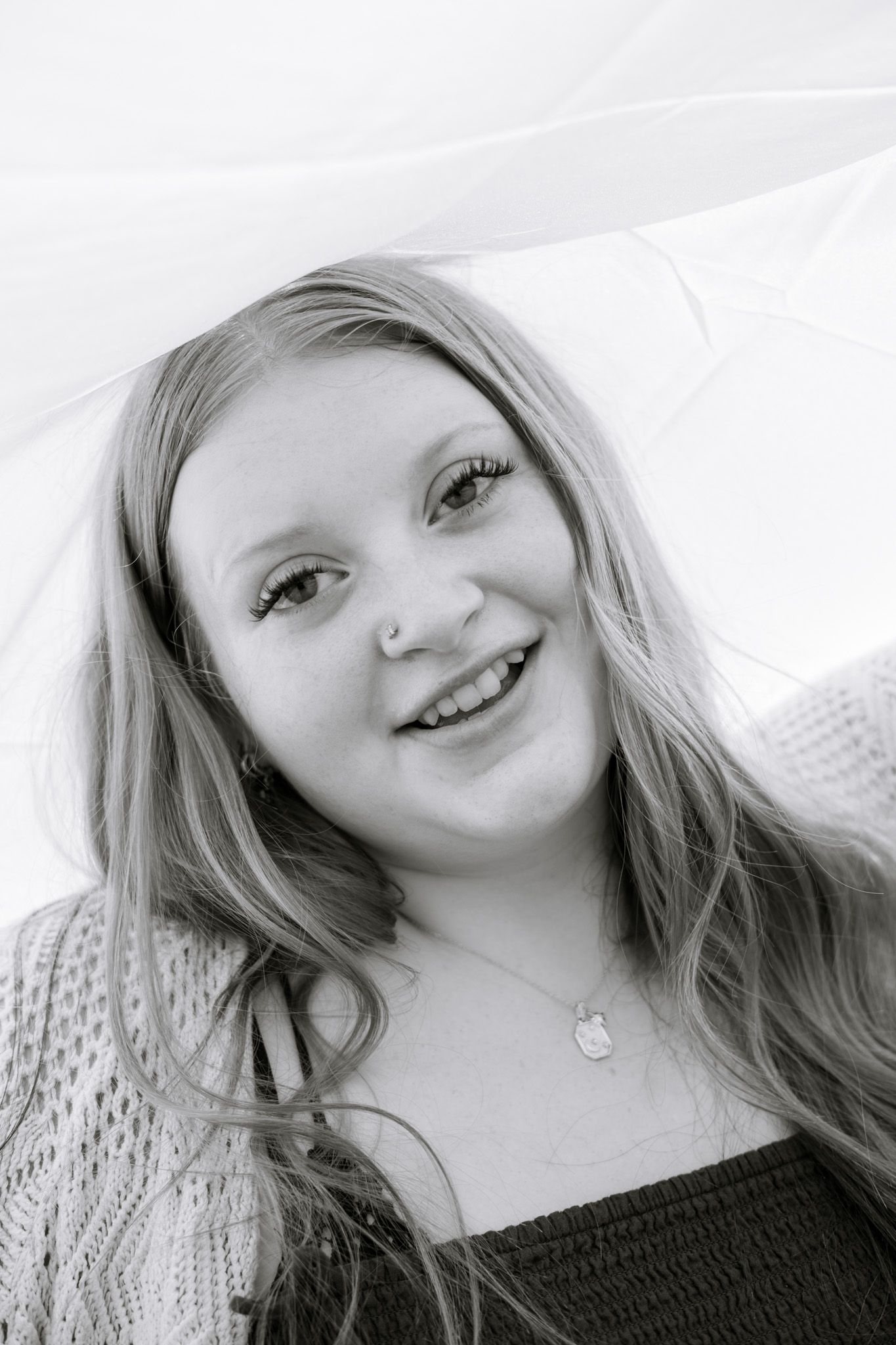 A young adult girl holds up a white sheet above her head and smiles. The image is in black and white.