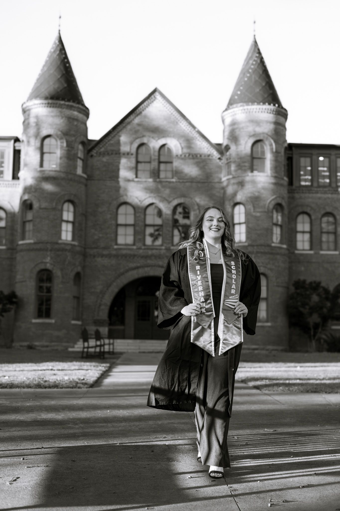 A young adult girl dressed in her regalia is walking in front of her historic campus building. The image is in black and white.