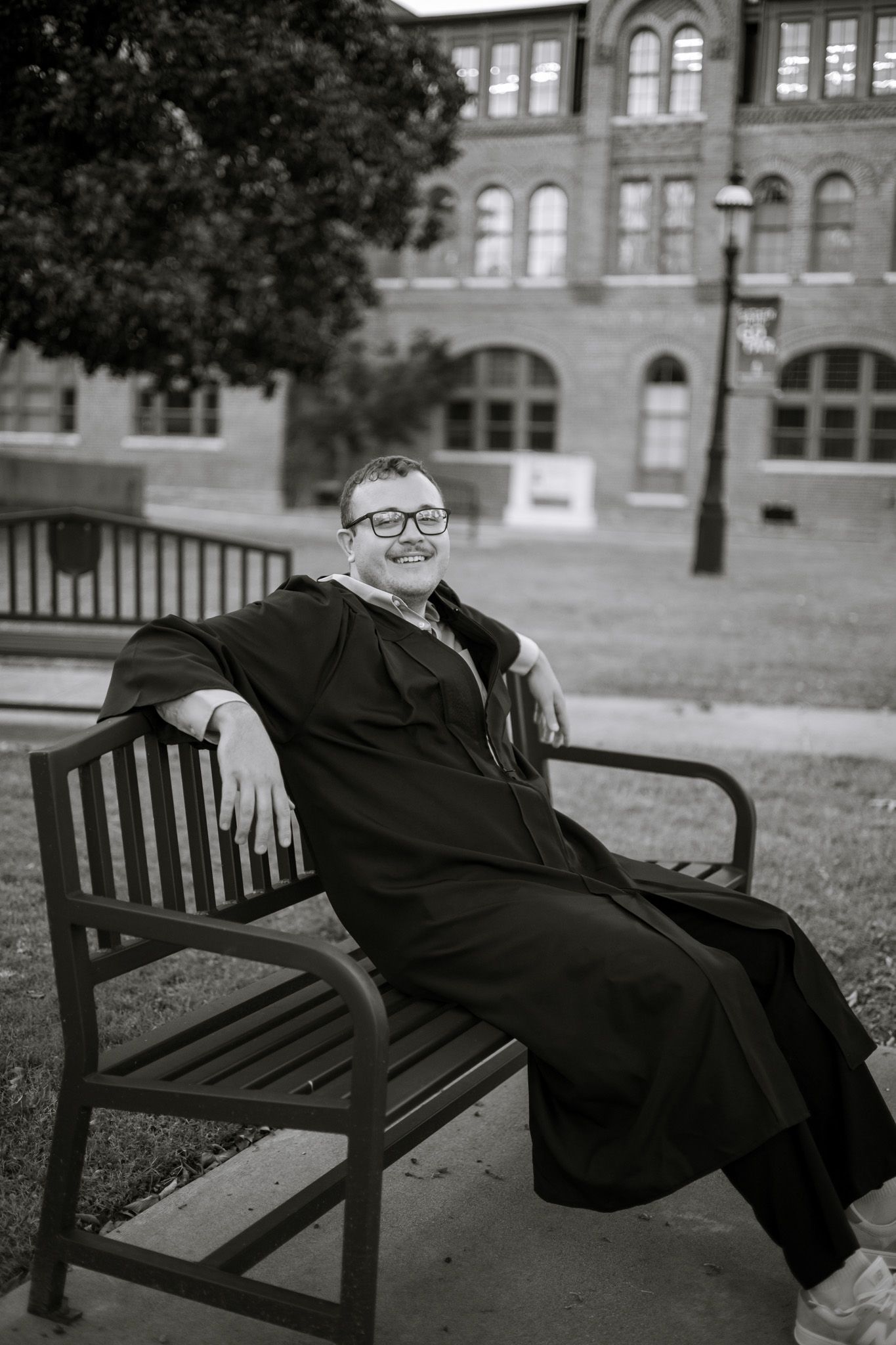 A young adult boy is resting against a bench in his graduation gown. He is smiling and looking away. The image is in black and white.