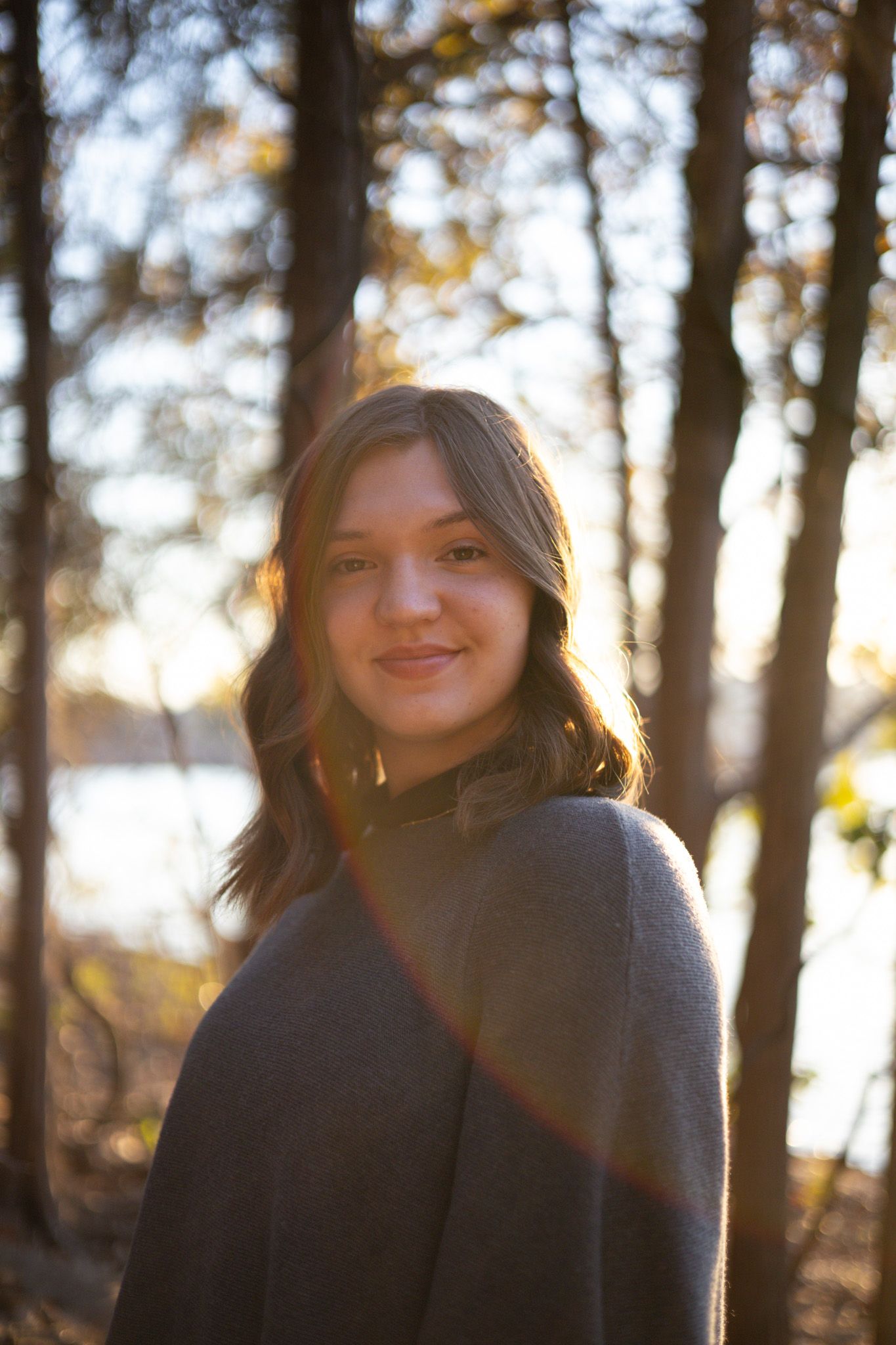 A young adult girl smiles in front of trees and a lake.