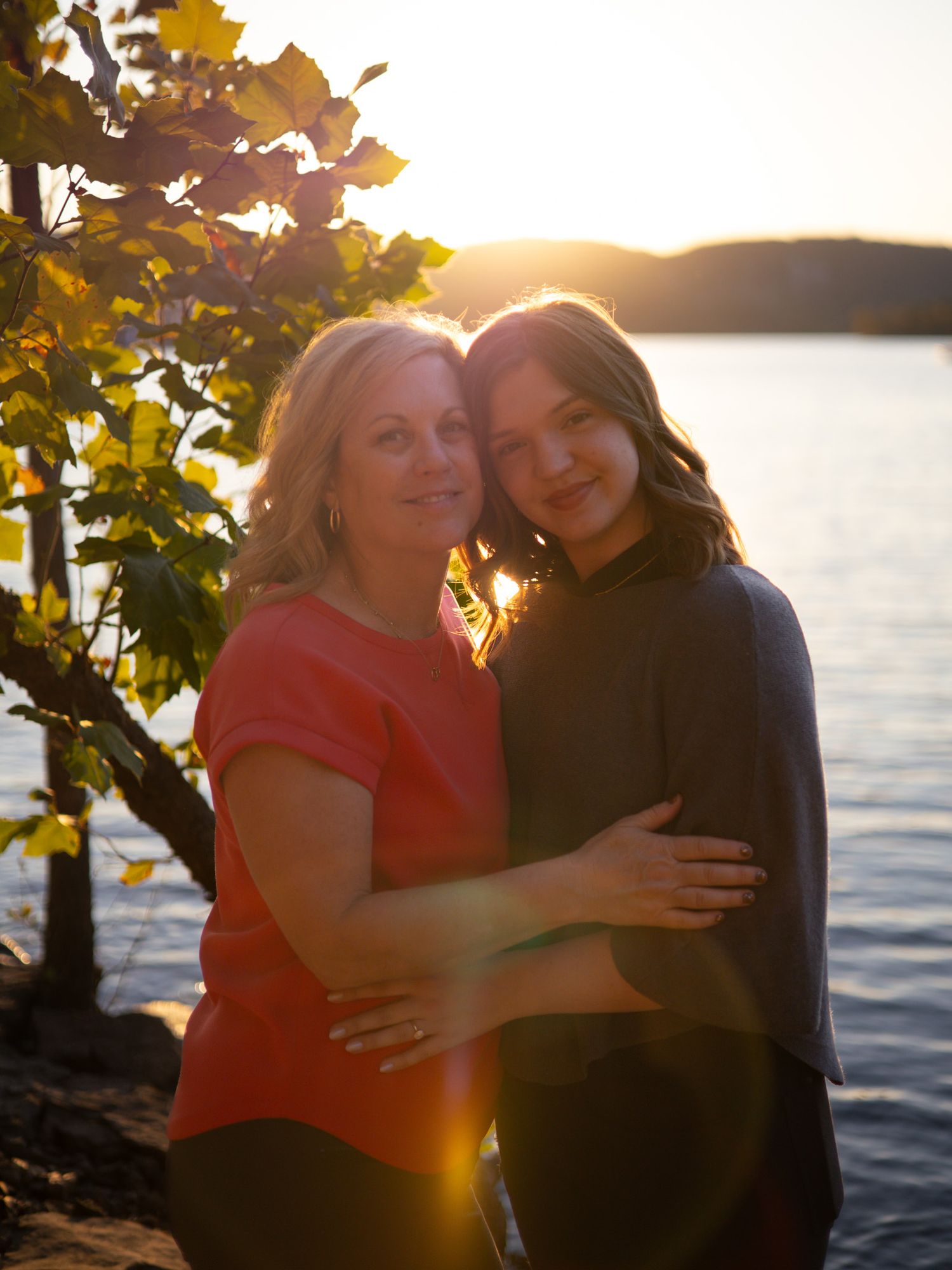 A mom and daughter hugging each other and facing the camera in front of a sunset on a lake.