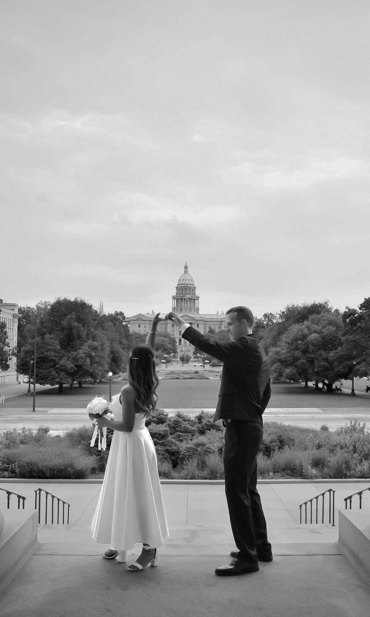 Black and white photo of a bride and groom holding hands on steps with a capitol building in the background.