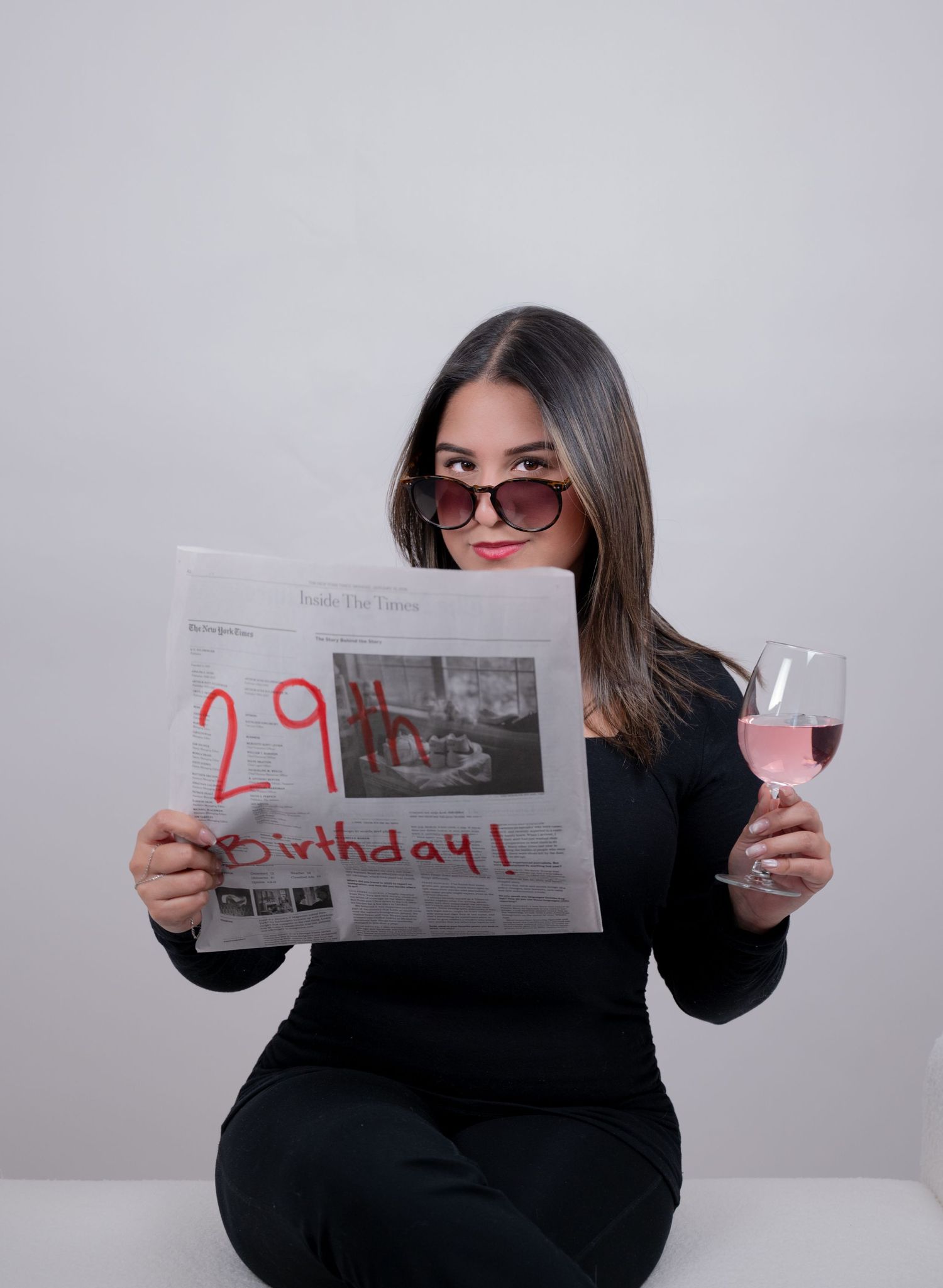 Woman in black outfit holding newspaper with 29th Birthday written in red while holding wine glass.