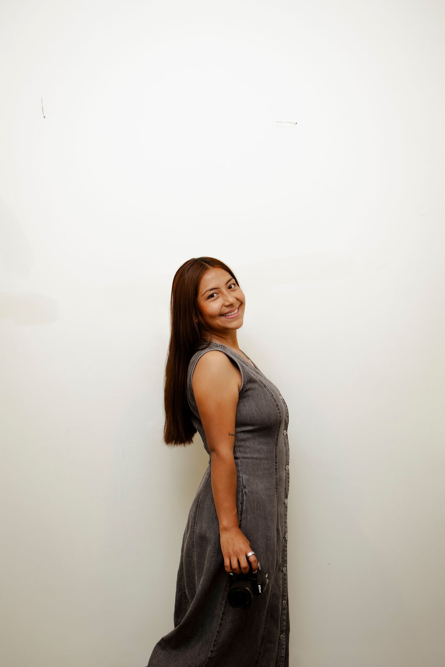 Smiling young Asian woman in gray sleeveless dress posing against a white wall holding a small purse.