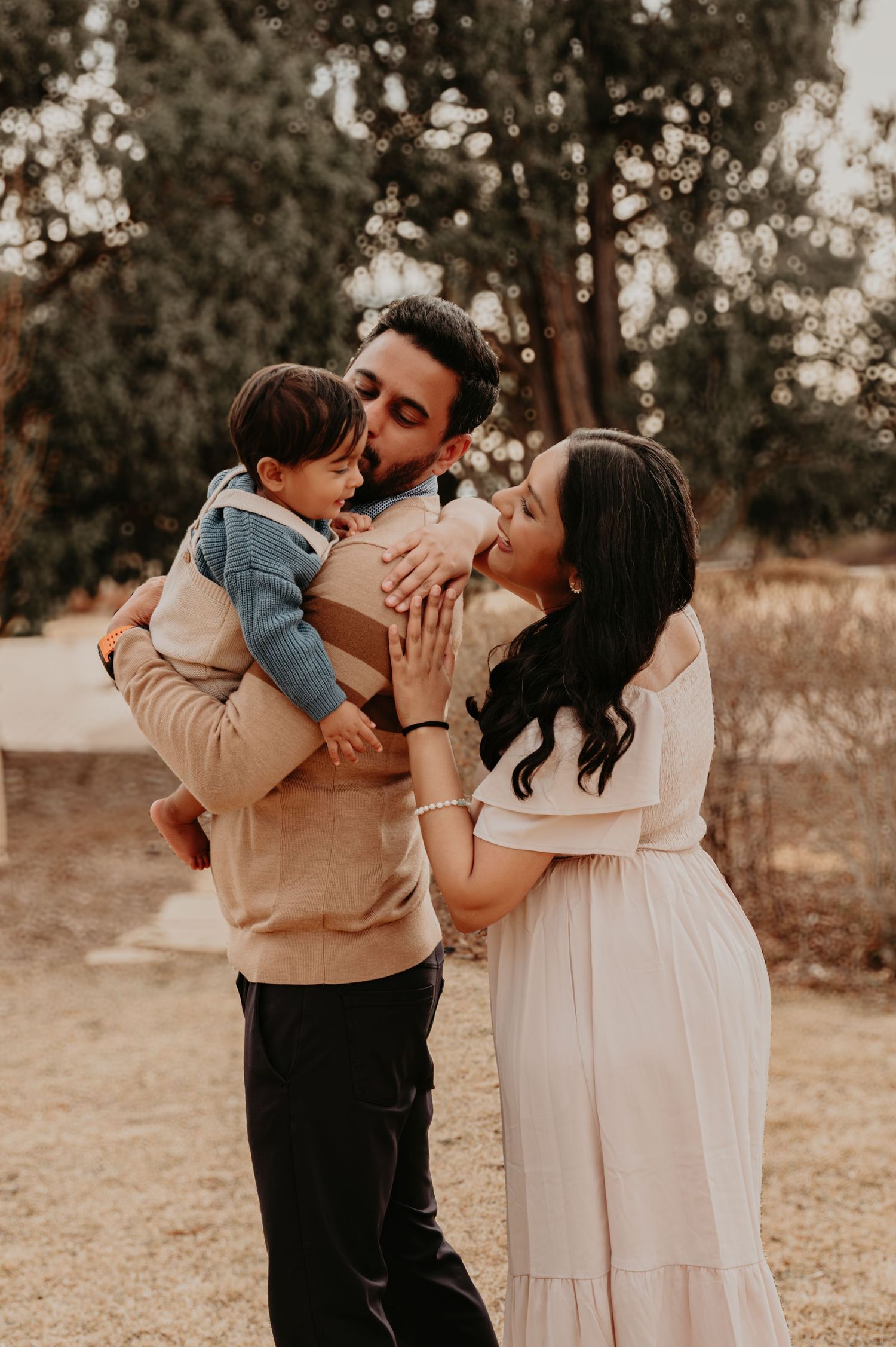 A couple shares a tender moment kissing their toddler outdoors, with the woman in a flowing pink dress showing a baby bump.