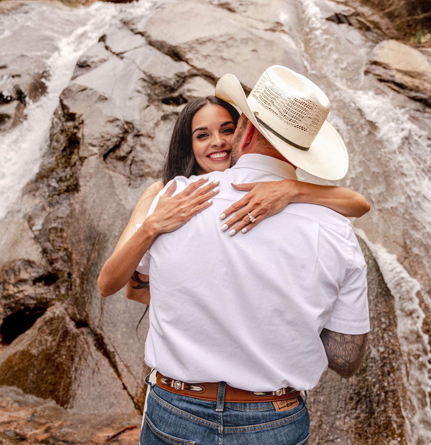 Couple embracing and smiling in front of a waterfall, woman showing engagement ring, man wearing cowboy hat.